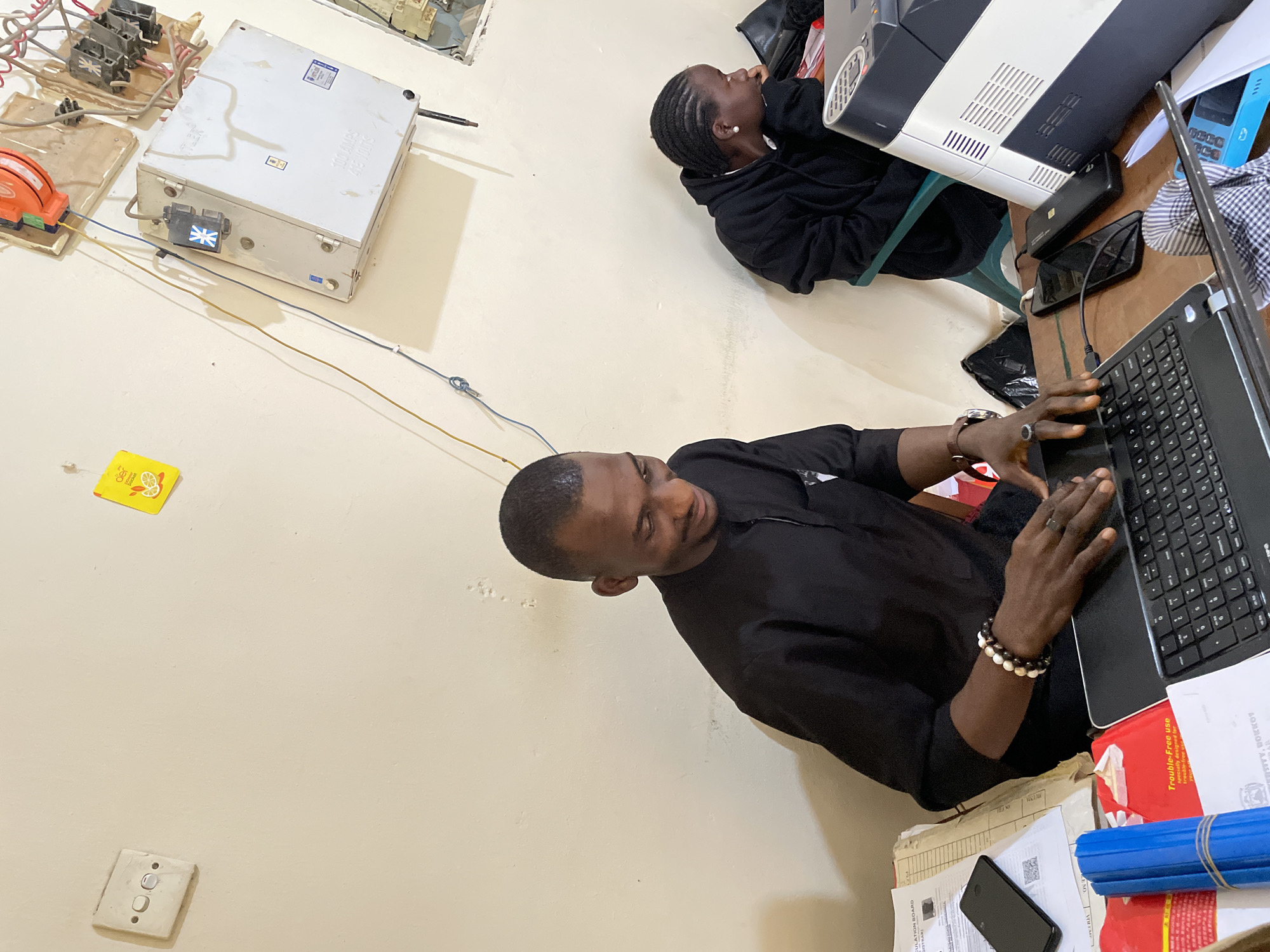 The image depicts an indoor scene, likely an office or administrative space in Durumi, Nigeria. A man dressed in a black traditional outfit is seated at a desk, looking down at a laptop. He is wearing bracelets and a ring. To his right, a woman in a black hoodie is also seated at a desk, her head resting on her hand, looking away from the camera.
On the desk in front of the man are papers, a smartphone, and a laptop. To his right, there is a printer and several other electronic devices and cables. The wall behind them is a light color and features electrical outlets, switches, and a distribution box with visible wiring. The overall environment appears functional and somewhat utilitarian.