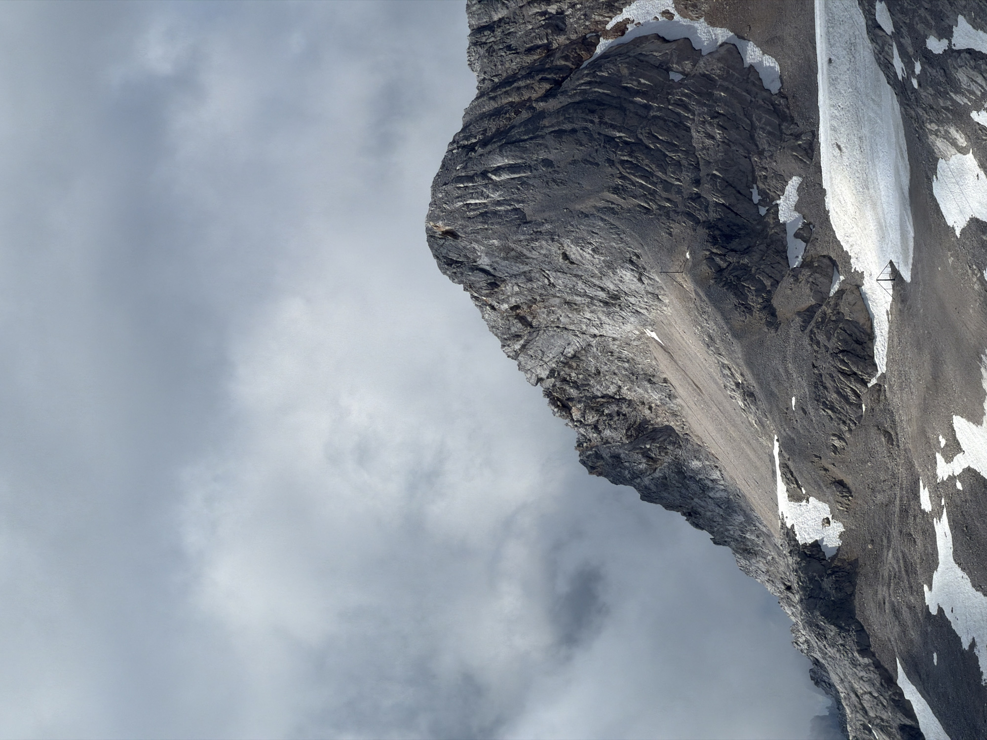The image depicts a dramatic mountain landscape with a prominent peak dominating the frame. The rock formations are rugged and textured, with visible layers and fissures. Patches of snow and ice are scattered across the lower slopes and in crevasses, suggesting a high altitude. The sky is overcast with thick, grey clouds that partially obscure the mountain's summit. In the foreground, there are signs of human presence, including what appear to be trail markers or small structures, as well as a fencing-like structure on a patch of snow. The scene conveys a sense of raw, natural beauty and the harshness of a mountainous environment. This is likely a view of the Zugspitze, the highest peak in Germany, located near Garmisch-Partenkirchen.