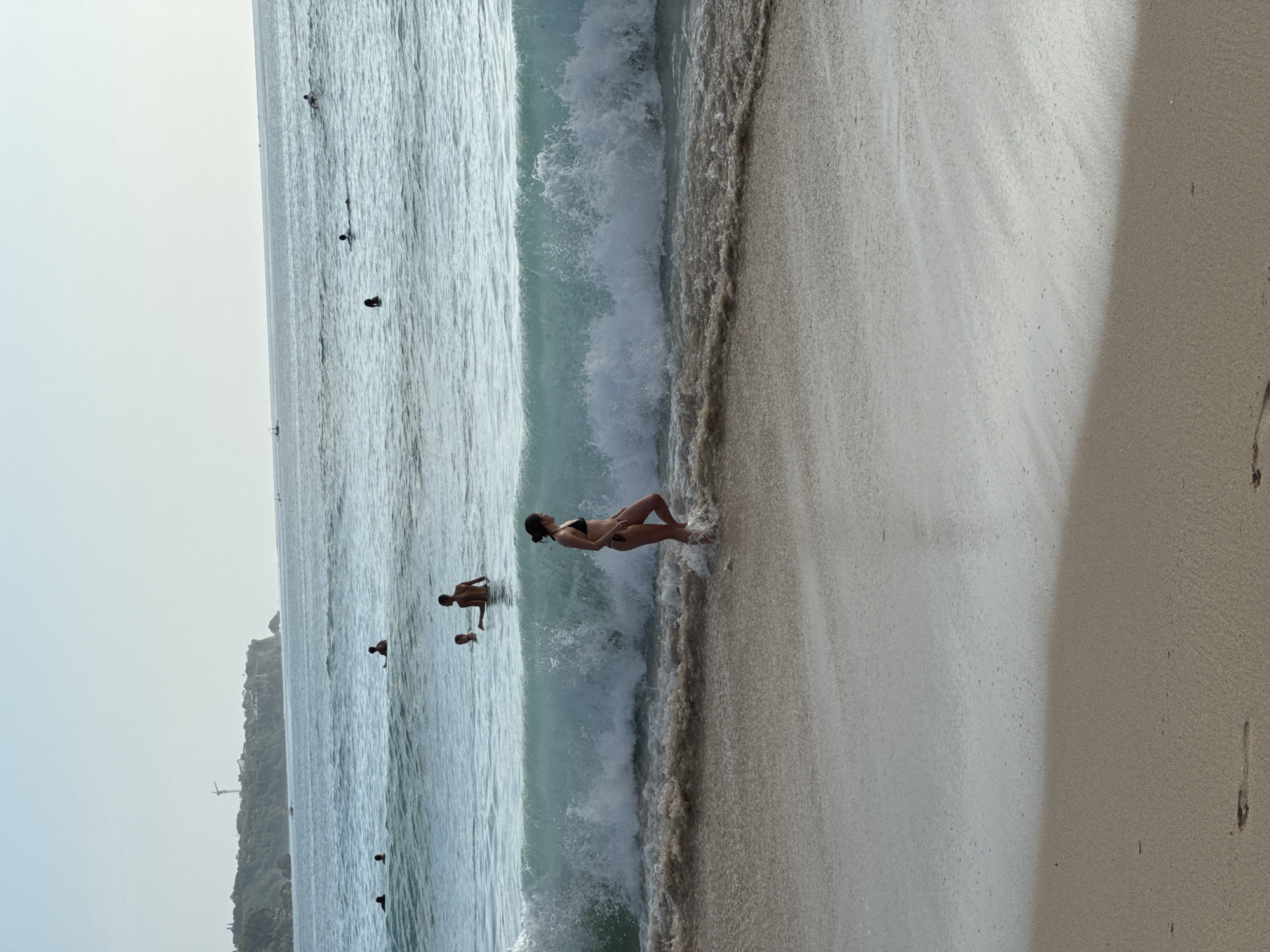 The image depicts a beach scene in Kuta Selatan, Indonesia. A woman in a black bikini lies on the sand near the shoreline, with waves washing over her. A man and a child are visible in the water nearby. In the distance, several other people are swimming in the ocean. A rocky cliffside and sparse structures are visible in the background. The beach itself is a mix of wet and dry sand. The sky is overcast, creating a hazy atmosphere.