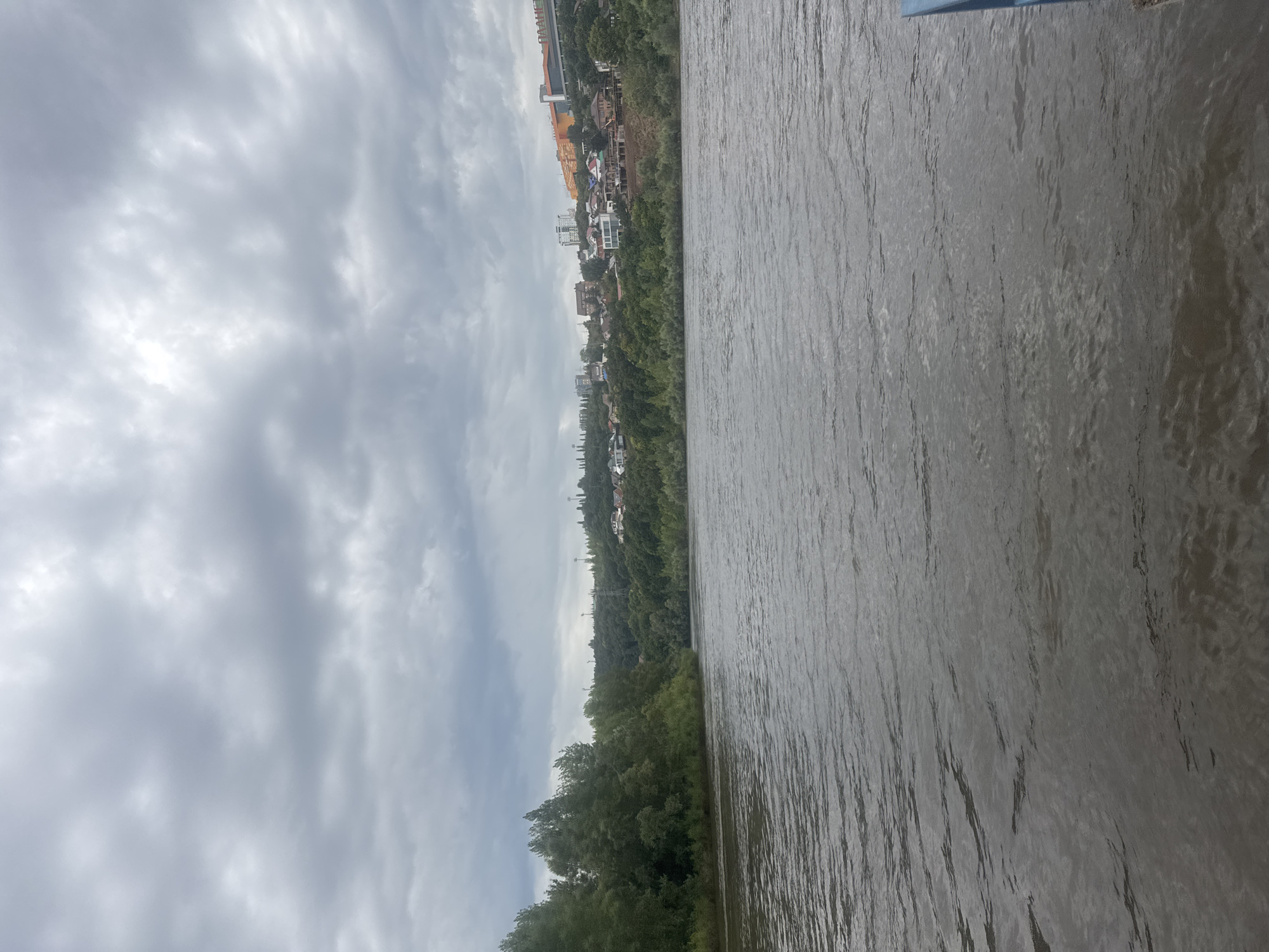The image depicts a wide, calm river flowing through a verdant landscape under a cloudy sky. The left bank is densely covered with trees, while the right bank features a residential area with houses, buildings, and some construction sites nestled amongst trees on a hillside. The river's surface shows ripples, indicating a gentle current. In the distance, power lines and towers are visible on the elevated terrain. The overall impression is of a tranquil, somewhat overcast day. The scene is consistent with views along the Belaya River in Ufa, Russia, known for its picturesque banks and residential development.