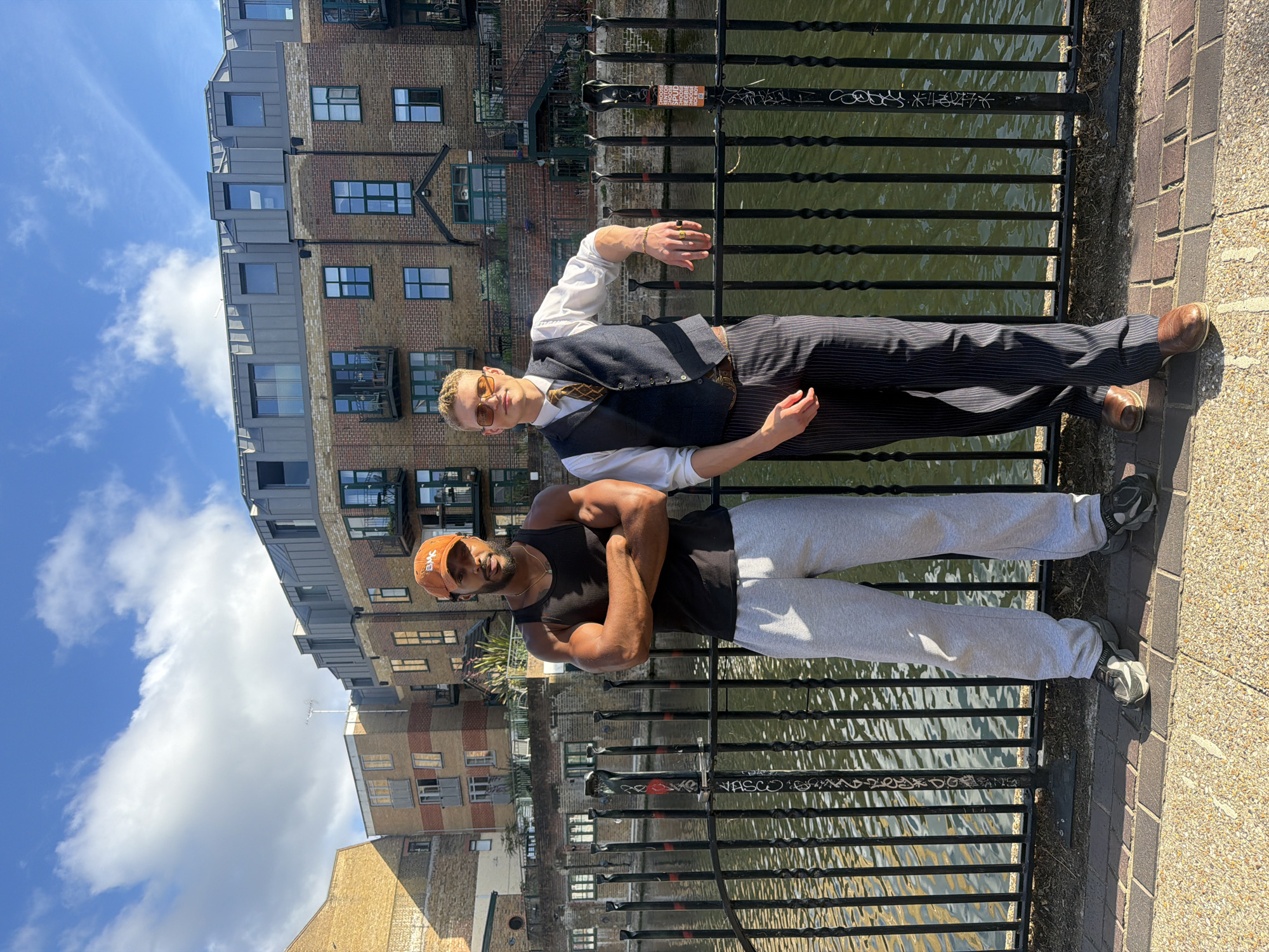 Two men stand by a canal in London. The man on the left is Black, wearing a black tank top, grey sweatpants, and a black baseball cap. He has his arms crossed. The man on the right is white, wearing a white button-down shirt, a dark waistcoat, pinstripe trousers, and brown shoes. He is wearing sunglasses and has his right hand resting on the canal railing. They are both facing the camera. The canal is to their left, with a black metal railing separating them from the water. Behind them is a multi-story building with brick and modern metal cladding, and a clear blue sky with scattered clouds above.