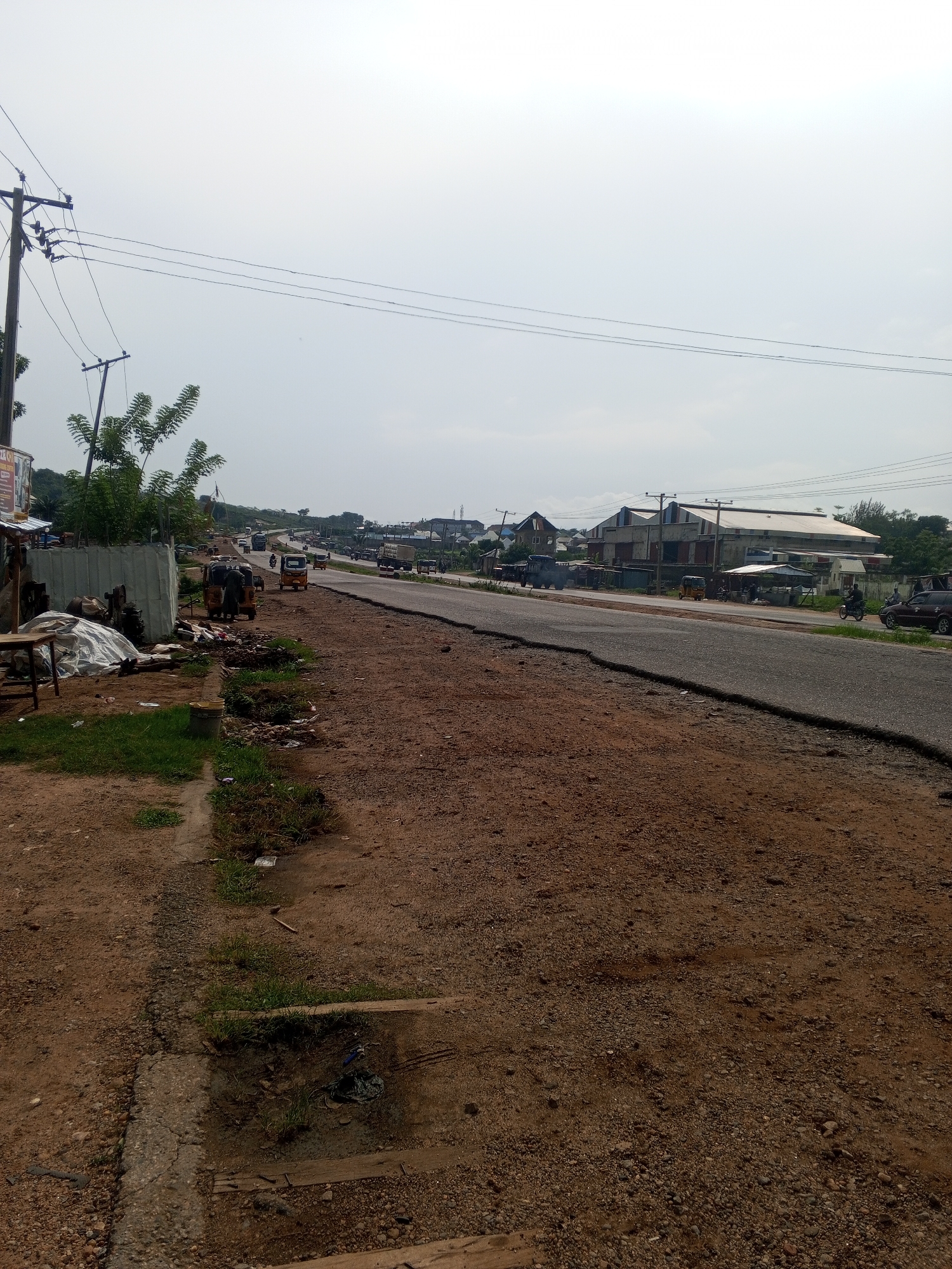 The image depicts a roadside scene in Lokoja, Nigeria, with a focus on the infrastructure and activity along a paved road. A dirt shoulder lines the road, littered with debris and patches of overgrown grass. On the left side of the road, a makeshift stall is set up with a wooden table, a tarpaulin-covered object, and what appears to be discarded machinery. Further down the road, several auto-rickshaws and motorcycles are visible, indicating local transportation. Buildings, including what looks like a large warehouse or industrial structure, line the opposite side of the road. Power lines stretch across the sky, supported by wooden utility poles. The sky is overcast, suggesting a possibly hazy or cloudy day. The overall impression is one of a developing urban environment with active commerce and transportation.