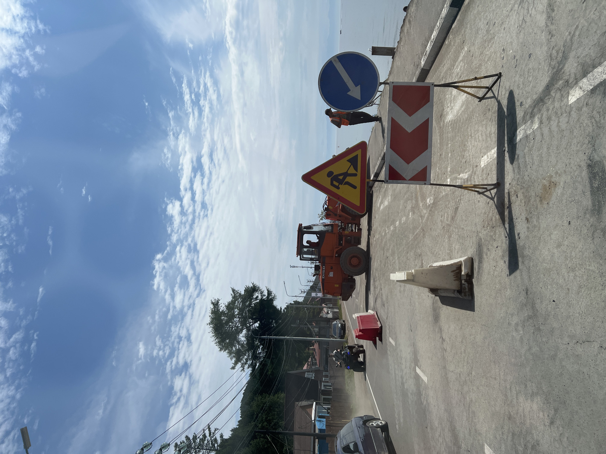 A construction scene is depicted along the shore of Lake Baikal in Listvyanka, Russia. An orange front-end loader with the logo "DISD" is present, indicating ongoing road work. Several traffic signs are visible, including a yellow triangular sign with a silhouette of a person digging, a circular blue sign with a white arrow pointing left, and a red and white chevron sign indicating a turn. Two construction workers, one wearing a high-visibility orange vest, are present near the signs and the loader. Temporary traffic barriers, some white plastic cones and some red plastic barriers, are placed on the asphalt road. An ATV with a rider is seen on the road, kicking up dust. A dark-colored car is parked to the left, and another car is further down the road. In the background, houses and trees line a hillside, and the calm waters of Lake Baikal are visible under a partly cloudy blue sky.