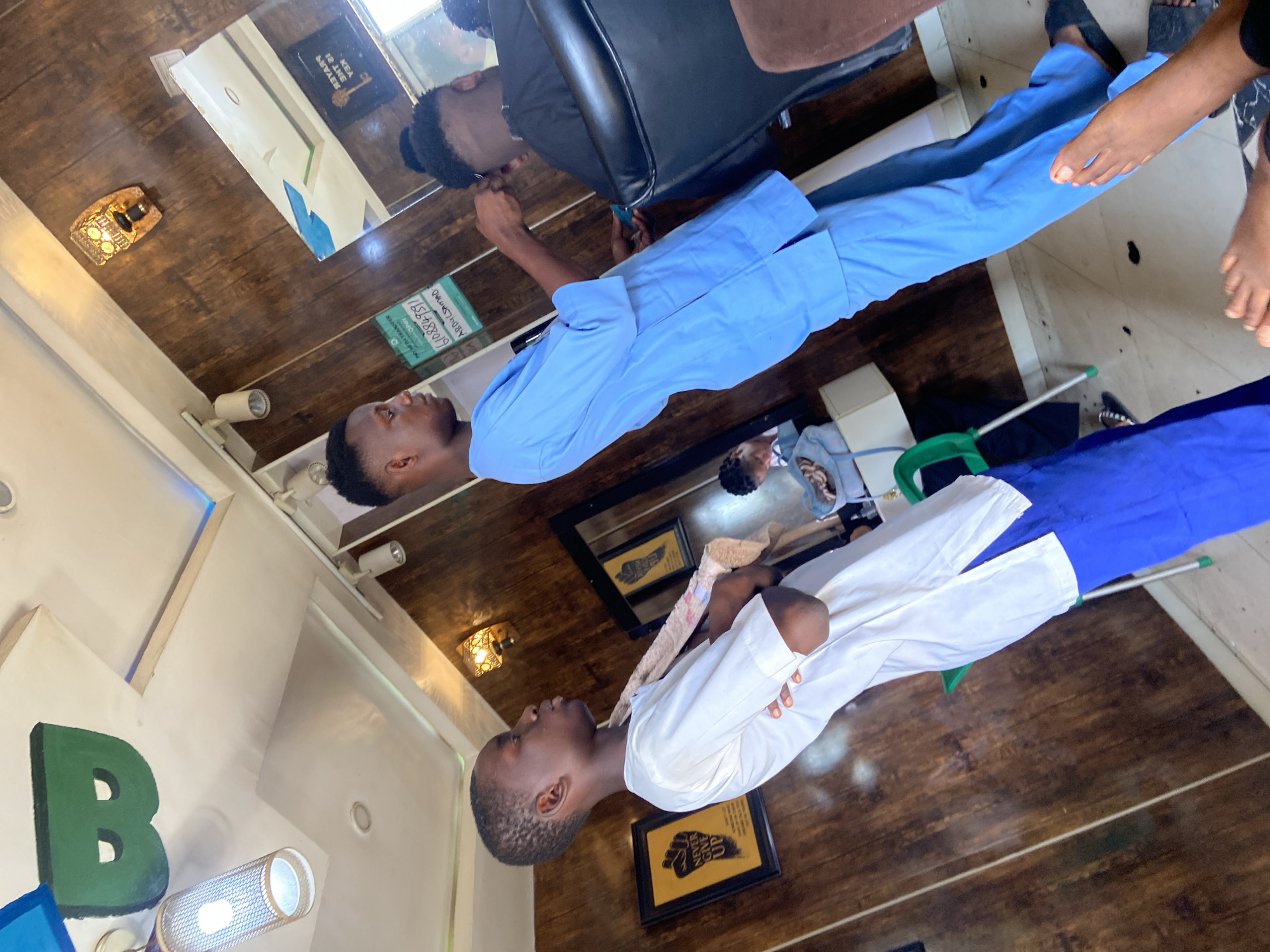 A brightly lit interior scene in what appears to be a barbershop in Lokoja, Nigeria. Two young Black men are the primary subjects. One man, seated in a black barber chair, is wearing a black shirt and has his hair styled in a small bun. He appears to be looking at a mobile phone. Reflected in a mirror are the man and a wall with a framed picture.
The second man, standing and wearing a white and blue outfit, is in the foreground and slightly out of focus. The wood-paneled floor of the shop is visible. A wall with a prominent green letter "B" is also visible. Other objects include light fixtures, a vacuum cleaner, and framed art. A pair of brown feet are partially visible in the upper right corner. The scene conveys a casual and social atmosphere.