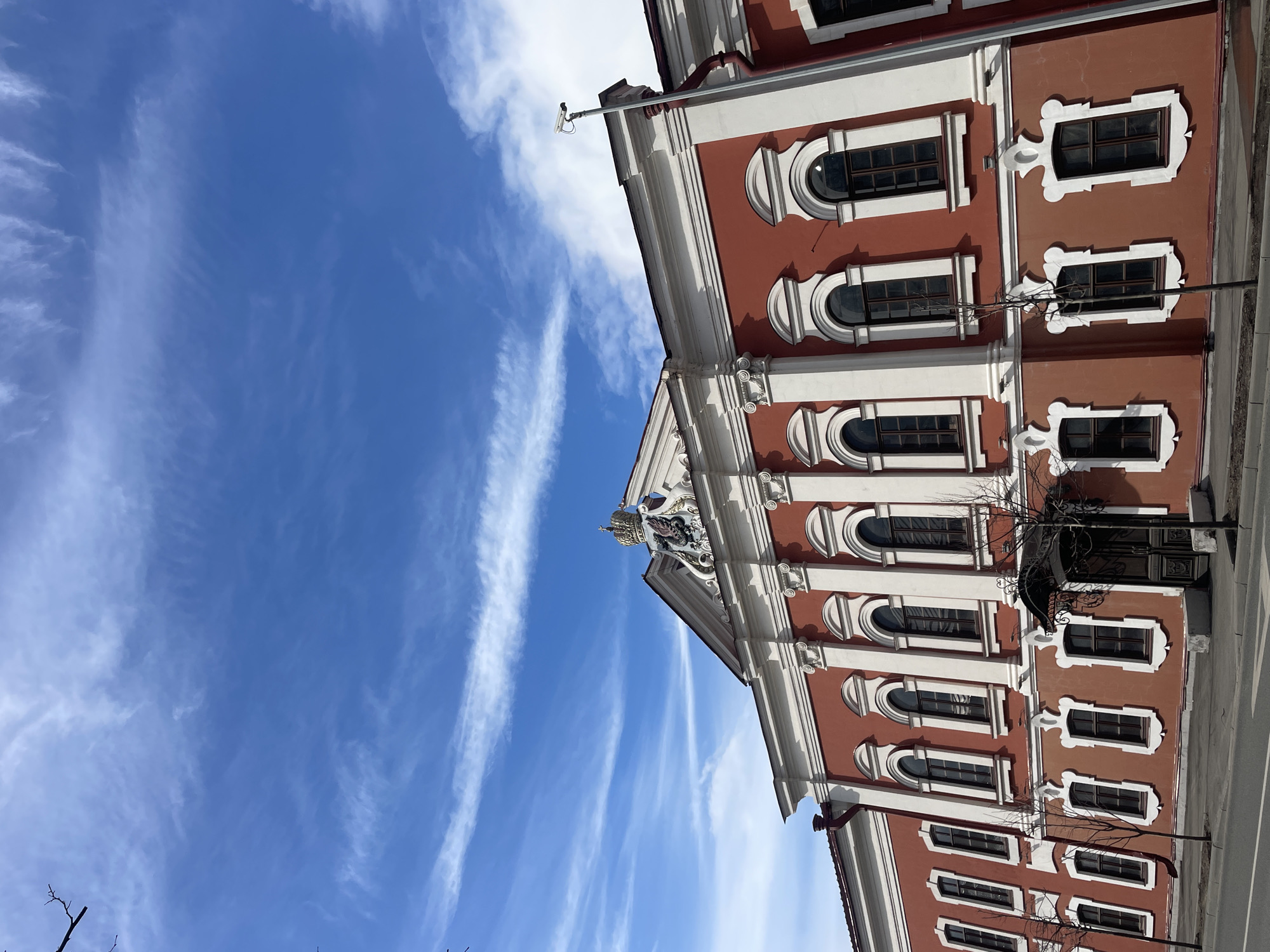 The image captures a grand, ornate building under a clear, bright blue sky adorned with wispy white clouds. The architecture features reddish-brown walls strikingly contrasted with elaborate white decorative elements. These include prominent cornices, pilasters, and intricate frames surrounding numerous windows. The windows are a mix of arched and rectangular shapes, all with highly decorative white surrounds, some with sculpted details.
At the apex of the building's facade, just below the roofline, an ornate crest or coat of arms is prominently displayed, topped with a golden crown, indicating historical or official significance. A white security camera on a pole is visible extending from the upper left portion of the building. Drainpipes are also discernible along the roofline. Bare tree branches are visible in the lower right and left corners, suggesting a season without foliage.
The sky dominates a significant portion of the composition, presenting a vibrant blue hue. Streaky, thin white clouds, possibly cirrus, are spread across the sky. The scene is brightly lit, indicative of a sunny daytime. No people or specific activities are visible. There is no discernible text within the image. The architectural style, with its classical and baroque influences, is characteristic of European cities, aligning with the given context of Kazan, Russia.