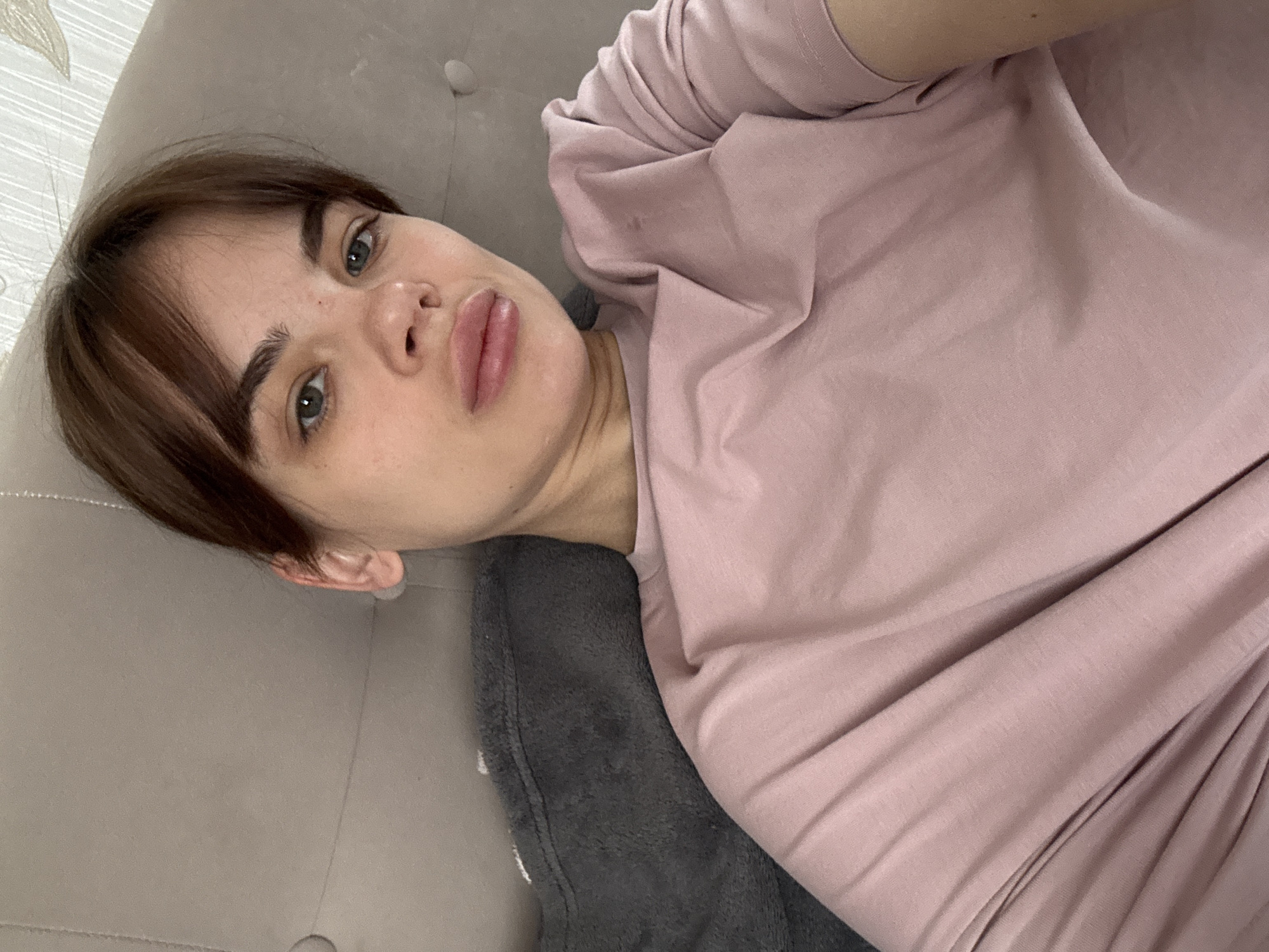 A woman with light skin, brown hair, and blue eyes is lying down on a gray couch. She is wearing a light pink shirt and resting on a gray pillow. The setting is indoors. The location is undetermined.