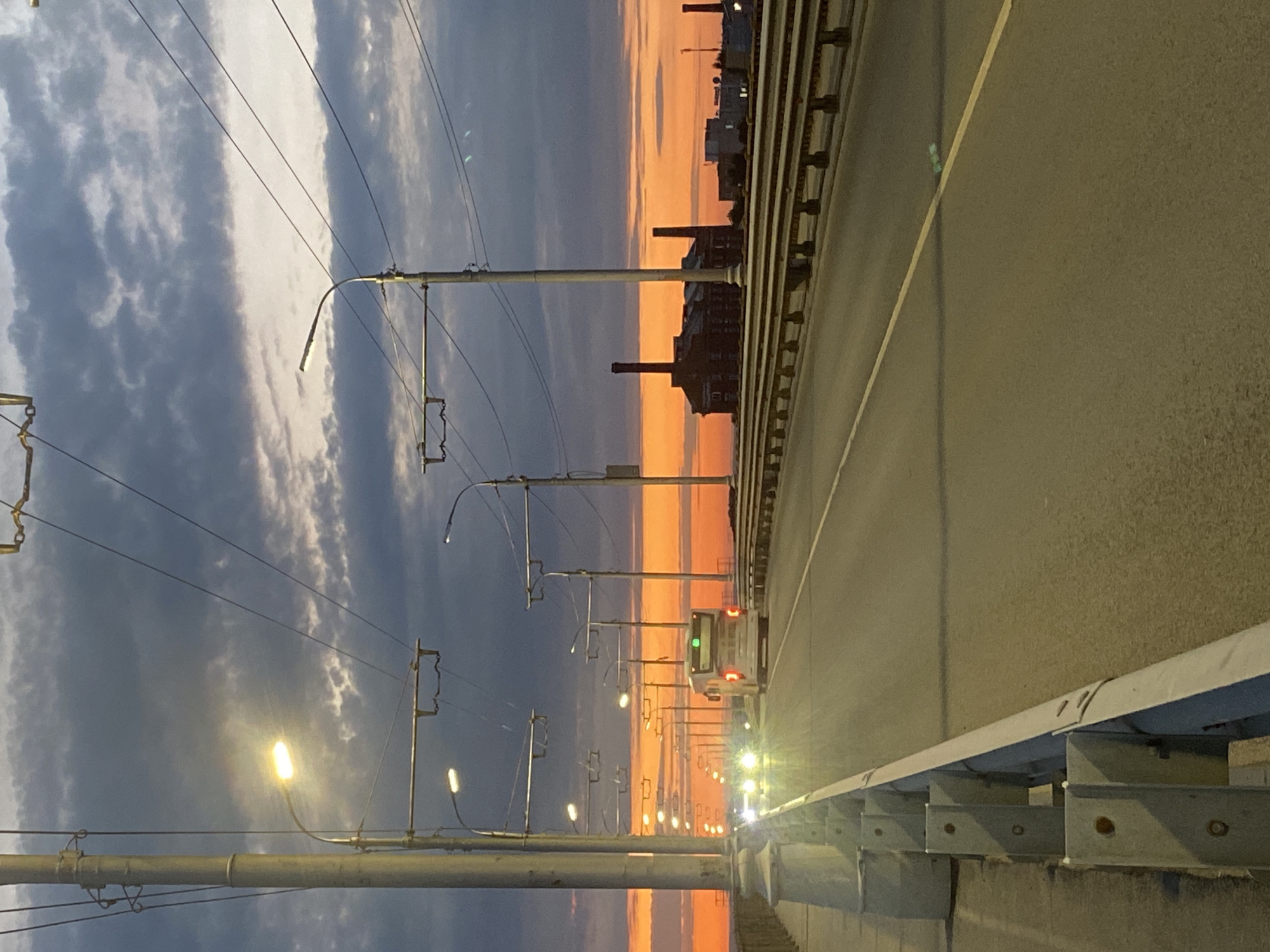 The image depicts a bridge over a road during a sunset. The sky is filled with dramatic clouds in shades of gray, dark blue, and vibrant orange. The sunset casts an orange glow across the horizon, illuminating industrial buildings with smokestacks in the distance. On the bridge, streetlights are illuminated, lining the road. A bus and two cars are visible on the road, moving away from the viewer. The bridge has a metal guardrail and concrete barriers along its edge. The scene appears to be in the city of Novokuybyshevsk, Russia, near the industrial zone.