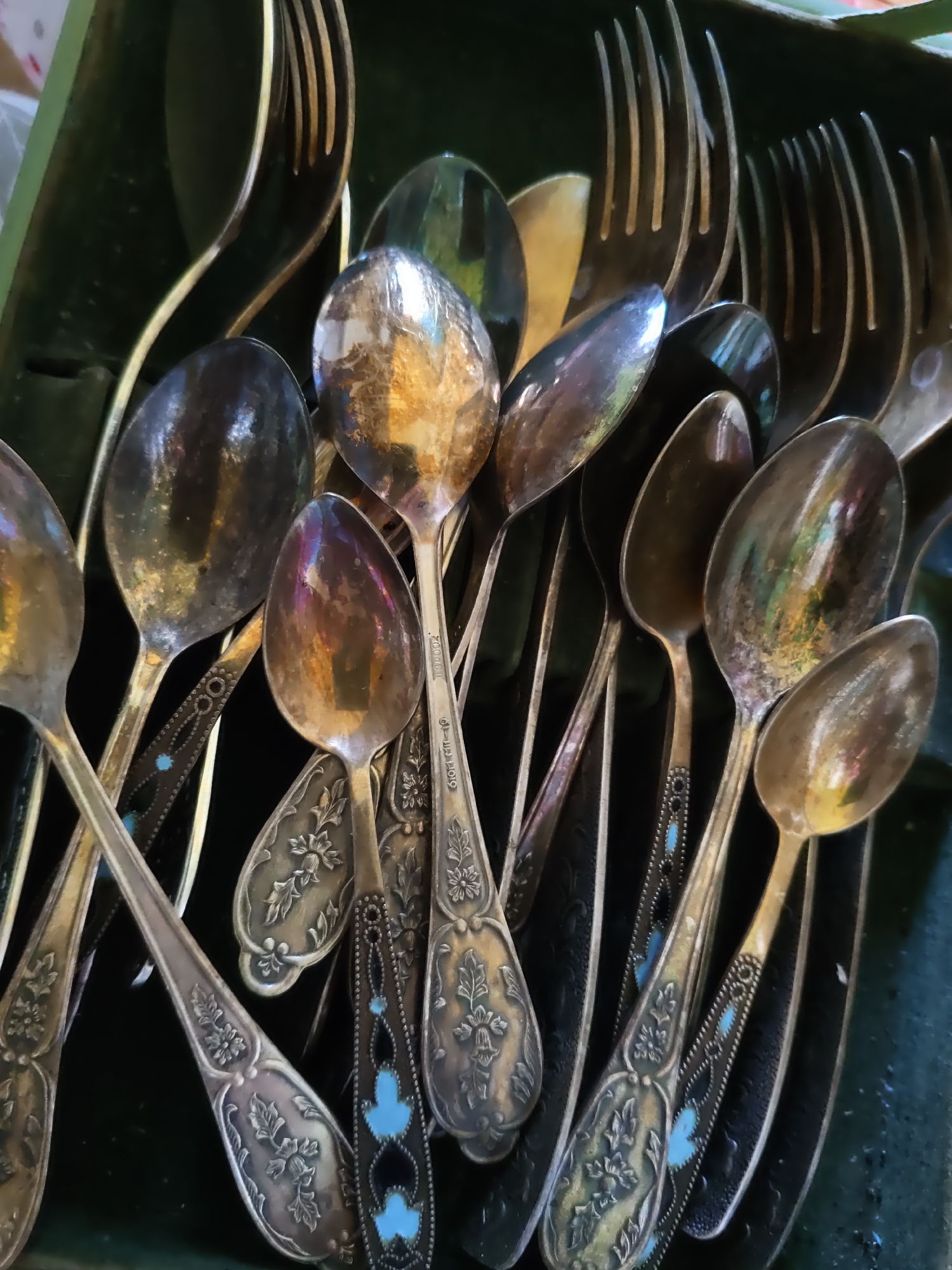 The image shows a close-up, overhead view of an assortment of antique silverware, primarily spoons and forks, lying in a dark green container. The silverware appears to be made of silver and exhibits tarnish and reflections from the surrounding light, giving them a varied iridescent sheen. Some of the spoons have intricate floral engravings on their handles, while others have contrasting patterns with small blue accents. The overall impression is one of historical items, possibly a collection or an antique store display. The context of Rostov-on-Don, Russia, is not visually represented in the image itself; there are no identifiable landmarks, people, or specific cultural markers to indicate this location.