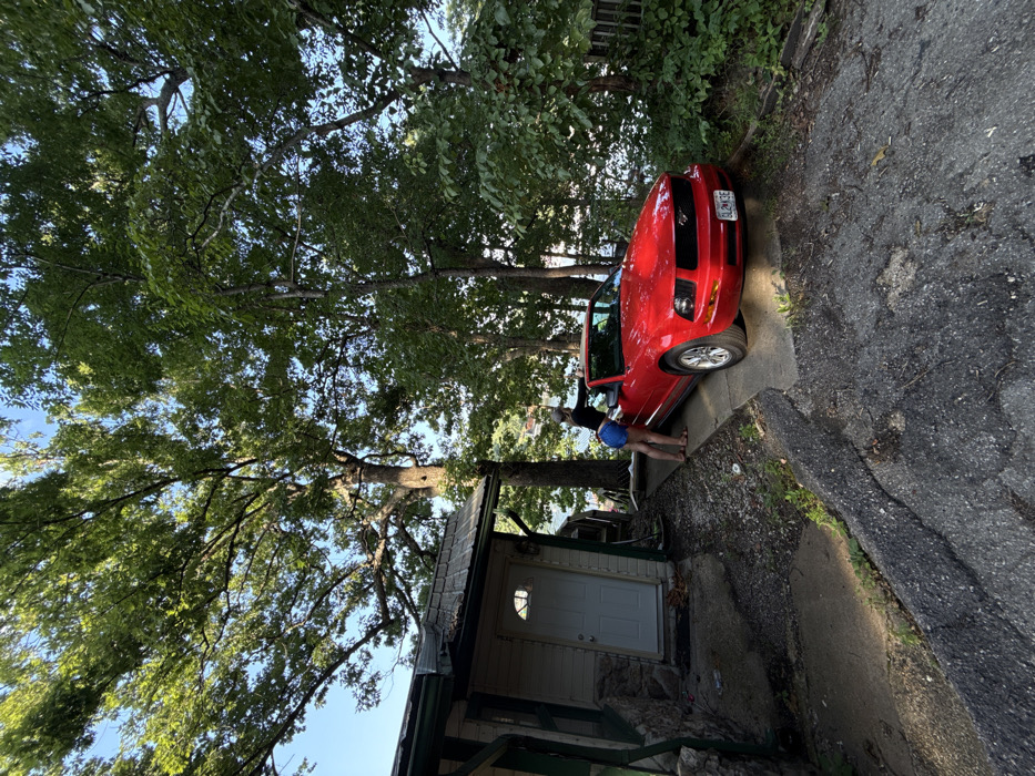 A red Ford Mustang convertible, likely from the 2005-2009 model generation, is parked on a concrete driveway in a wooded residential area of Osage Beach, United States. The car's soft top is retracted, and its front license plate reads "QLM3QE." A female individual, dressed in dark shorts, a dark top, and light-colored flip-flops, stands leaning into the open driver's side of the vehicle, with her back mostly to the viewer. To the immediate left of the car, a portion of a light-colored house with a stone foundation and a white front door is visible. The scene is dominated by numerous large trees with green foliage. A body of water is visible in the background through the trees, indicative of a lakeside property. The sky is clear, and the ground transitions from worn, dark asphalt in the foreground to a cleaner concrete slab beneath the car.