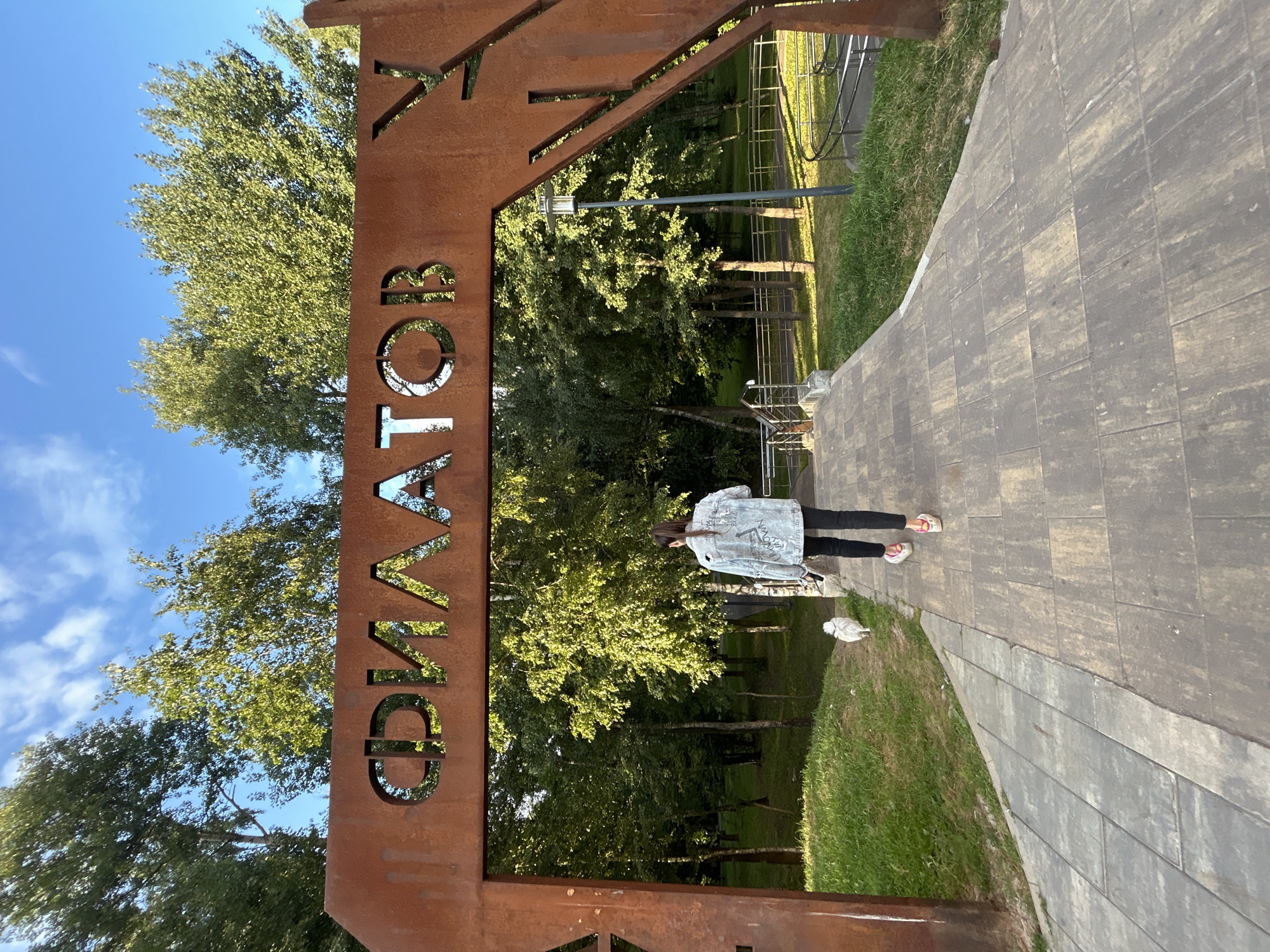 In the city of Moskovskiy, Russia, a person with their back to the camera walks away from the viewer on a paved pathway. They are wearing a light-colored denim jacket with white lettering, black pants, and pink sneakers. A small, fluffy white dog walks slightly ahead and to the left of the person. The pathway leads through a rustic, rusted metal archway with Cyrillic letters cut out, which appear to spell "ФИЛАТОВ". Beyond the arch, a park landscape is visible with numerous green trees, grass, a lamppost, and a metal railing along a sloped path. The sky is blue with scattered white clouds. The scene is outdoors during daylight.