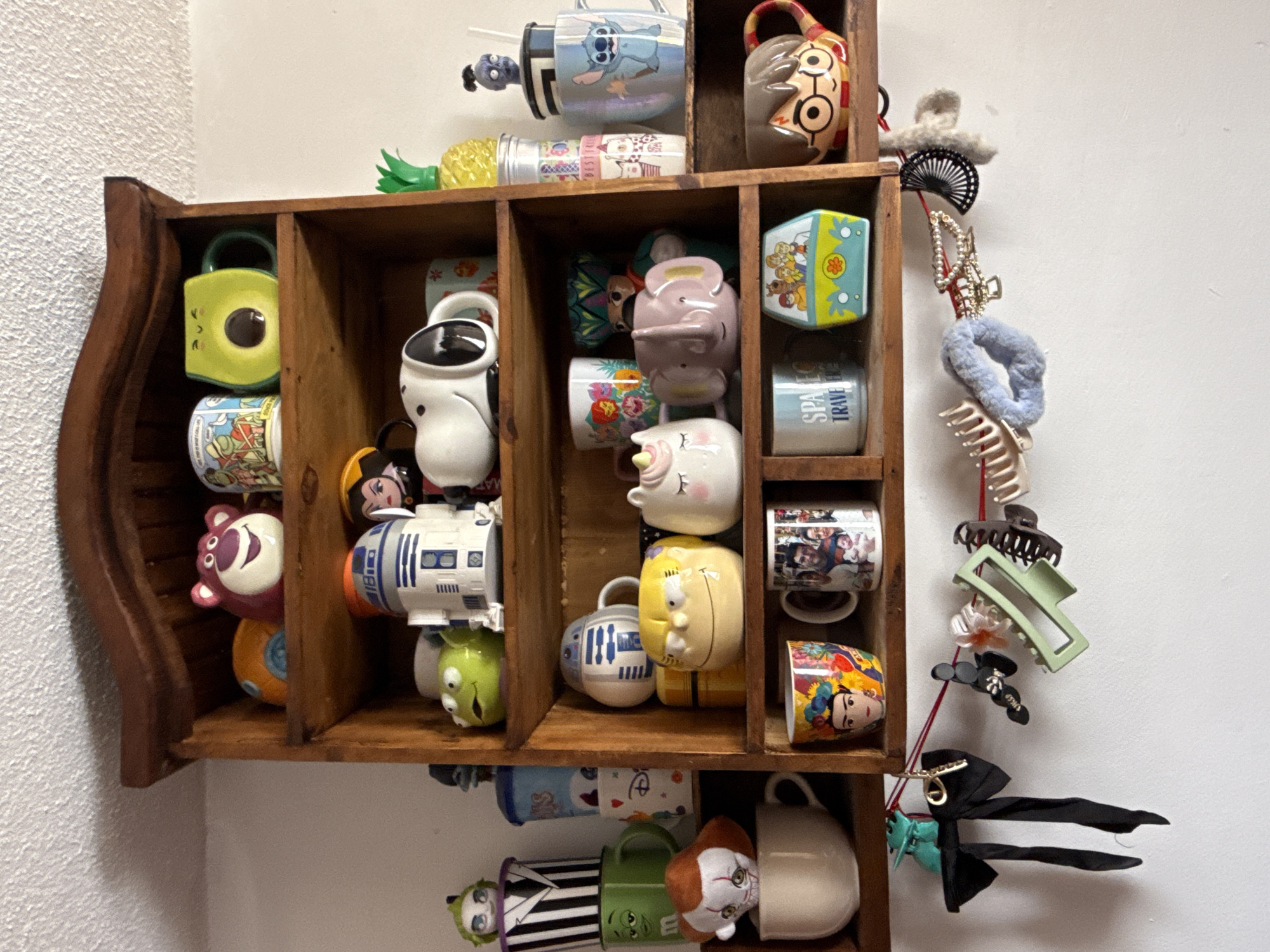 A wooden shelf is mounted on a white wall, filled with novelty mugs of various cartoon characters and other objects. The shelf has three columns and four rows. The mugs include characters from Disney, Star Wars, and other popular culture franchises. Various hair accessories, including clips and scrunchies, are hanging from the right side of the shelf. The mugs and accessories are arranged in a decorative, seemingly personal manner. The specific location within Puebla, Mexico, cannot be determined from the image.