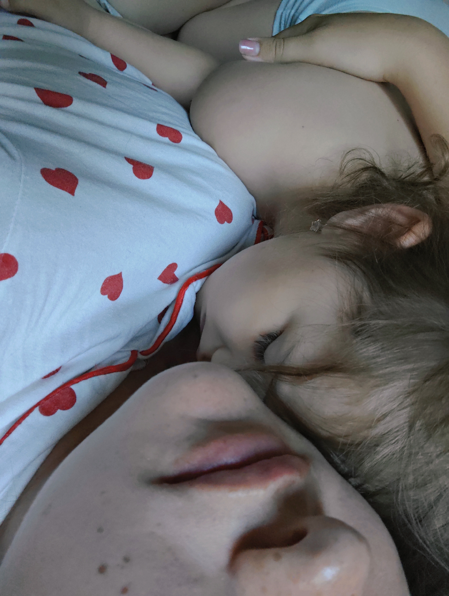 A close-up photograph captures a sleeping child resting on an adult, presumably a parent. The child's face, framed by light brown hair, is turned towards the adult's face. The adult's face is visible in the foreground, showing a portion of the nose and mouth. Both faces appear light-skinned. They are lying on a bed covered with a white sheet patterned with red hearts, and the adult is wearing light blue clothes. No location in Chaikovsky, Russia, is discernible from the image.