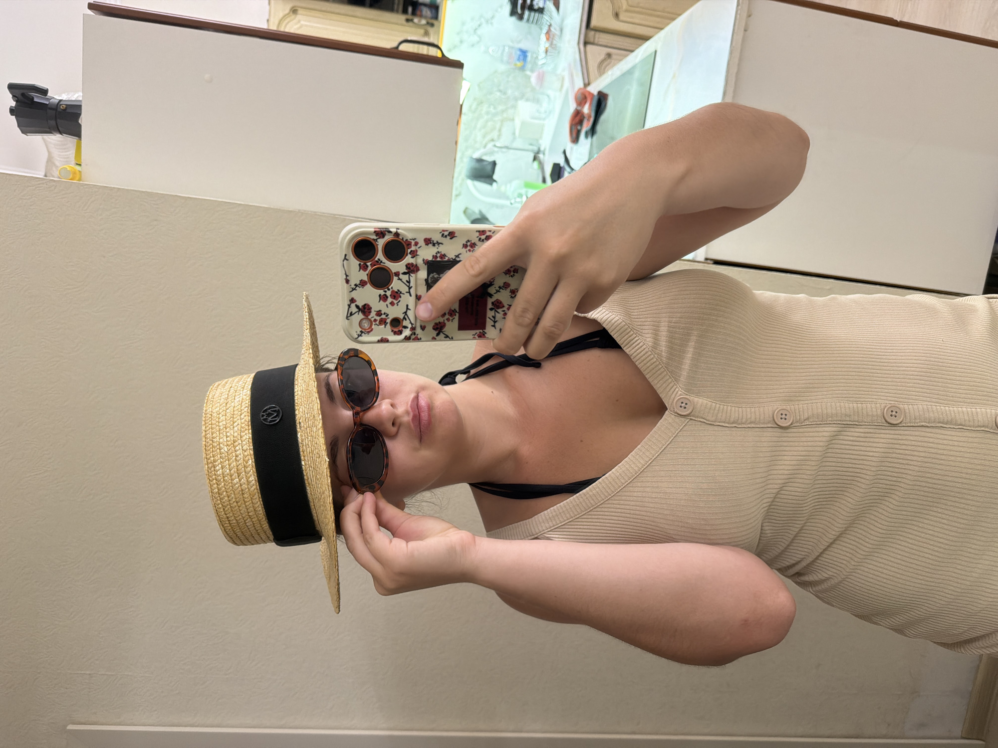 A young woman is taking a selfie in a kitchen. She is wearing a straw hat with a black band, sunglasses, and a beige ribbed dress with buttons. She is holding a phone with a floral case in her right hand. The kitchen appears to be in a residential setting, with white cabinets, a countertop, and some appliances visible in the background. The image does not provide any specific details that would allow it to be located in Sochi, Russia.