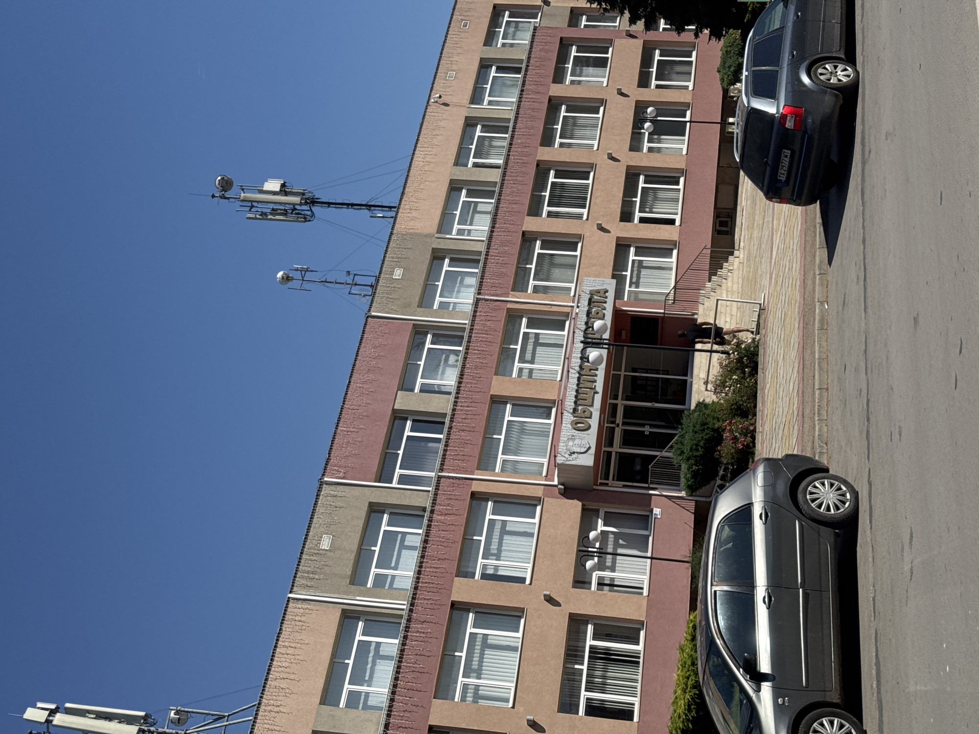 In Shabla, Bulgaria, a three-story municipal building with a light brown, pink, and grey facade stands under a clear blue sky. The building features multiple windows with white frames on each level, and some windows have vertical blinds. A sign above the entrance reads "ОБЩИНА ШАБЛА" (Municipality of Shabla). A person wearing a black t-shirt and shorts is walking up a set of stairs to the building's entrance. Two cars are parked on the street in front of the building; a dark grey hatchback is on the left, and a dark grey station wagon is on the right, displaying a license plate with "TX 2977 MT". Several communication towers with antennas are visible on the roof of the building and in the background. Green bushes and some flowering plants are situated in front of the building.