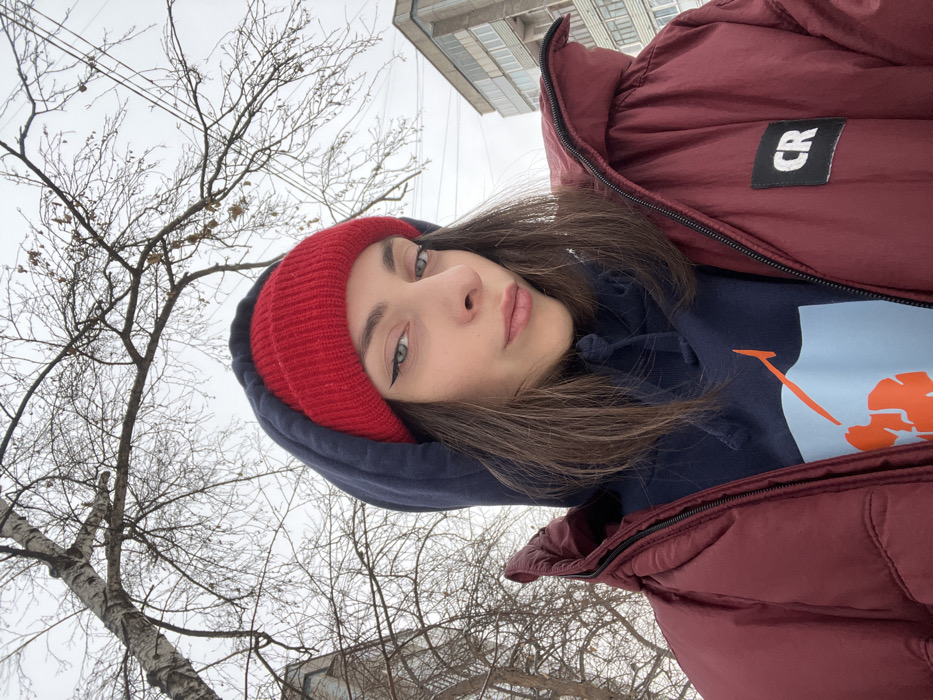 A woman takes a selfie outdoors. She wears a red beanie, a dark blue hoodie, and a maroon puffer jacket with a "CR" patch. Bare trees fill the left of the frame against a gray sky, with a building partially visible. The woman has fair skin, dark hair, and light-colored eyes with dark eyeliner.