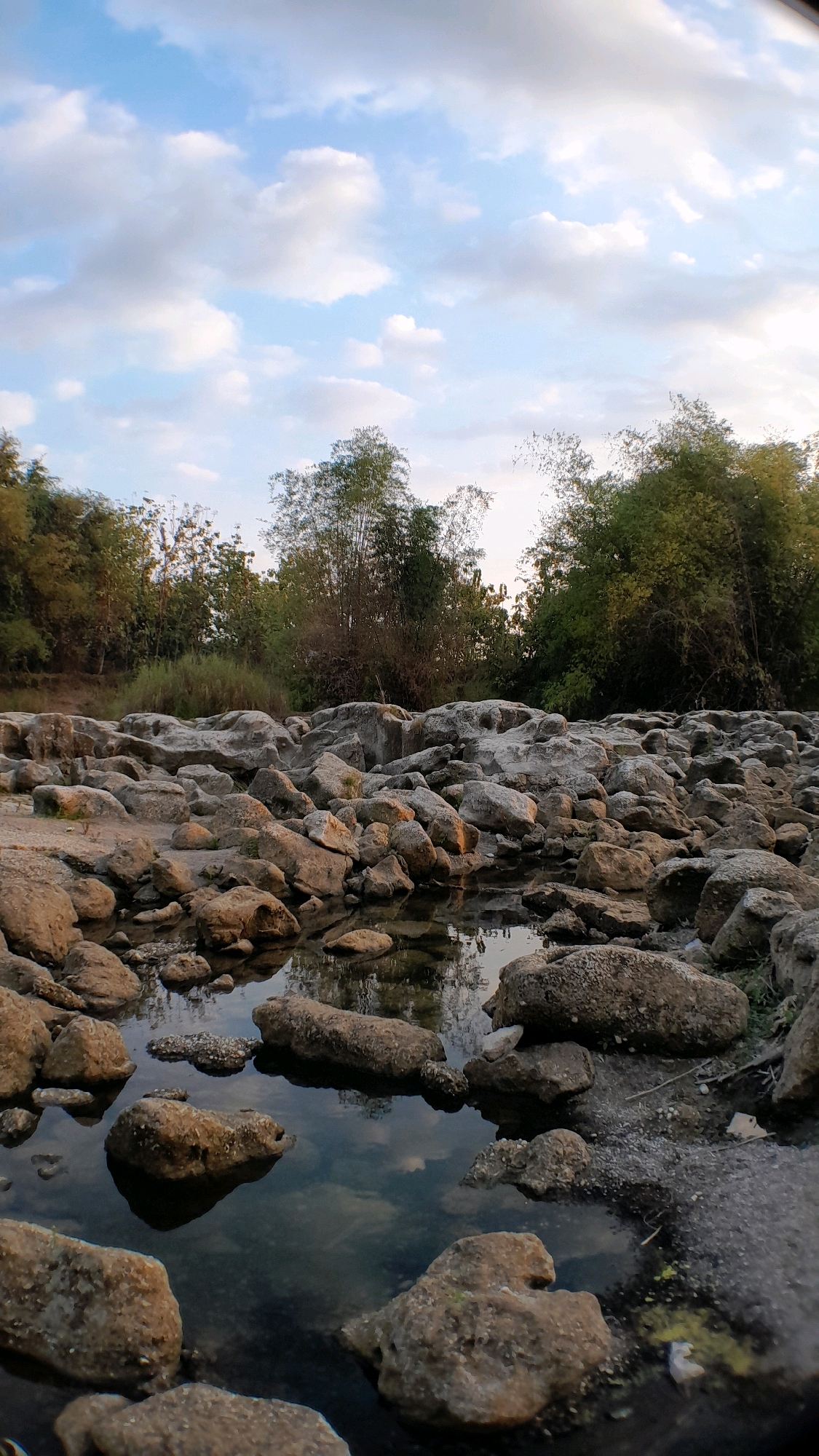 This image depicts a rocky riverbed with scattered rocks and shallow water reflecting the sky. Tall trees, including bamboo, line the background, suggesting a natural, possibly rural or park-like setting. The sky is partly cloudy with a mix of blue sky and white clouds. The lighting suggests it is either early morning or late afternoon. The absence of people or any human activity indicates a tranquil scene. The location is identified as Ketuwan, Indonesia.