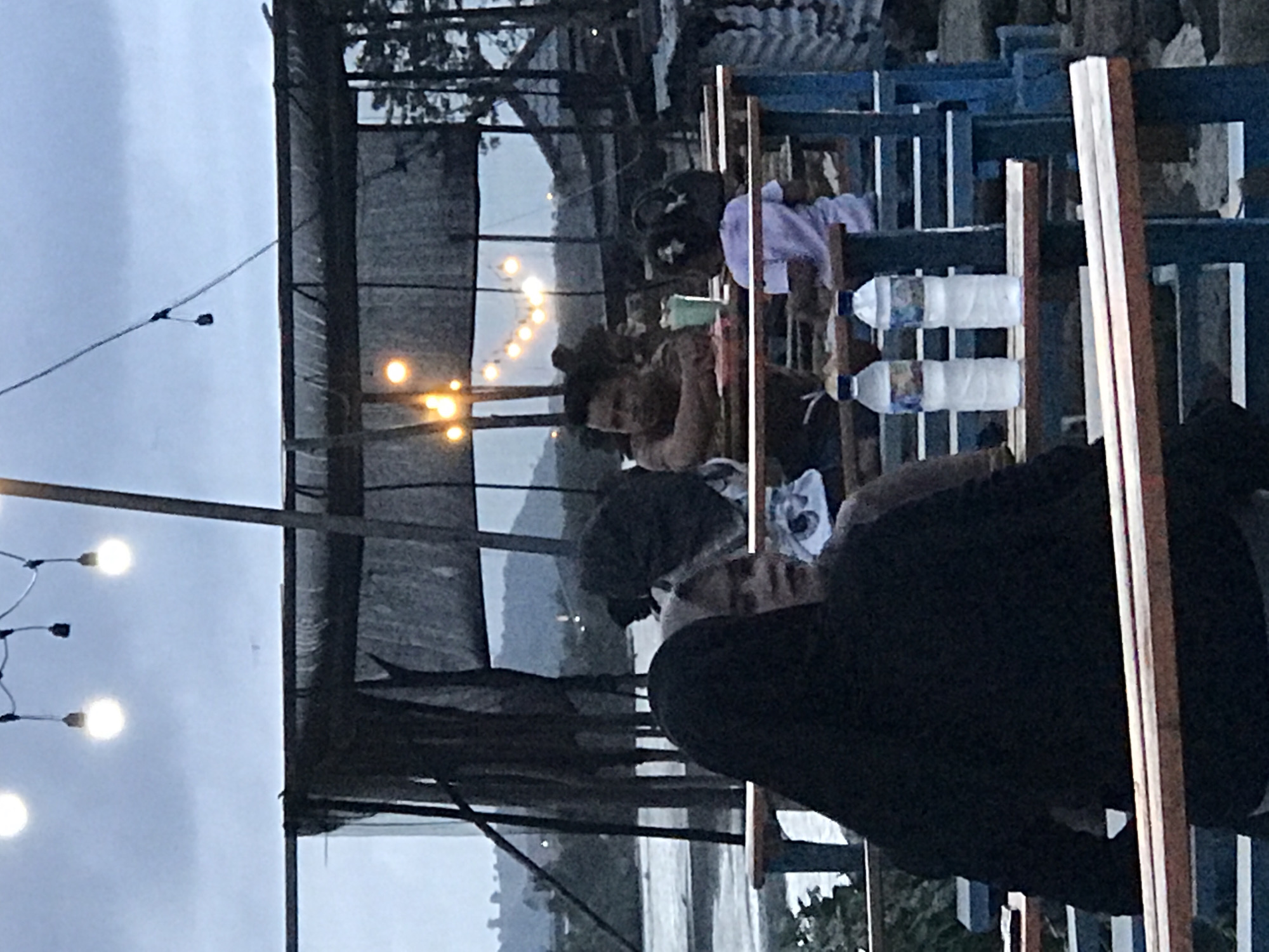 The image shows a group of people sitting at wooden tables, possibly at an outdoor restaurant or gathering place, in Lhokseumawe, Indonesia. The setting appears to be near a body of water, possibly the sea, with land visible in the distance. The lighting suggests it is dusk or evening, with strung lights illuminating the scene. People are engaged in conversation or looking at their phones, and bottled water is visible on the table. The structures around them are simple, with corrugated metal roofing and open-air construction.