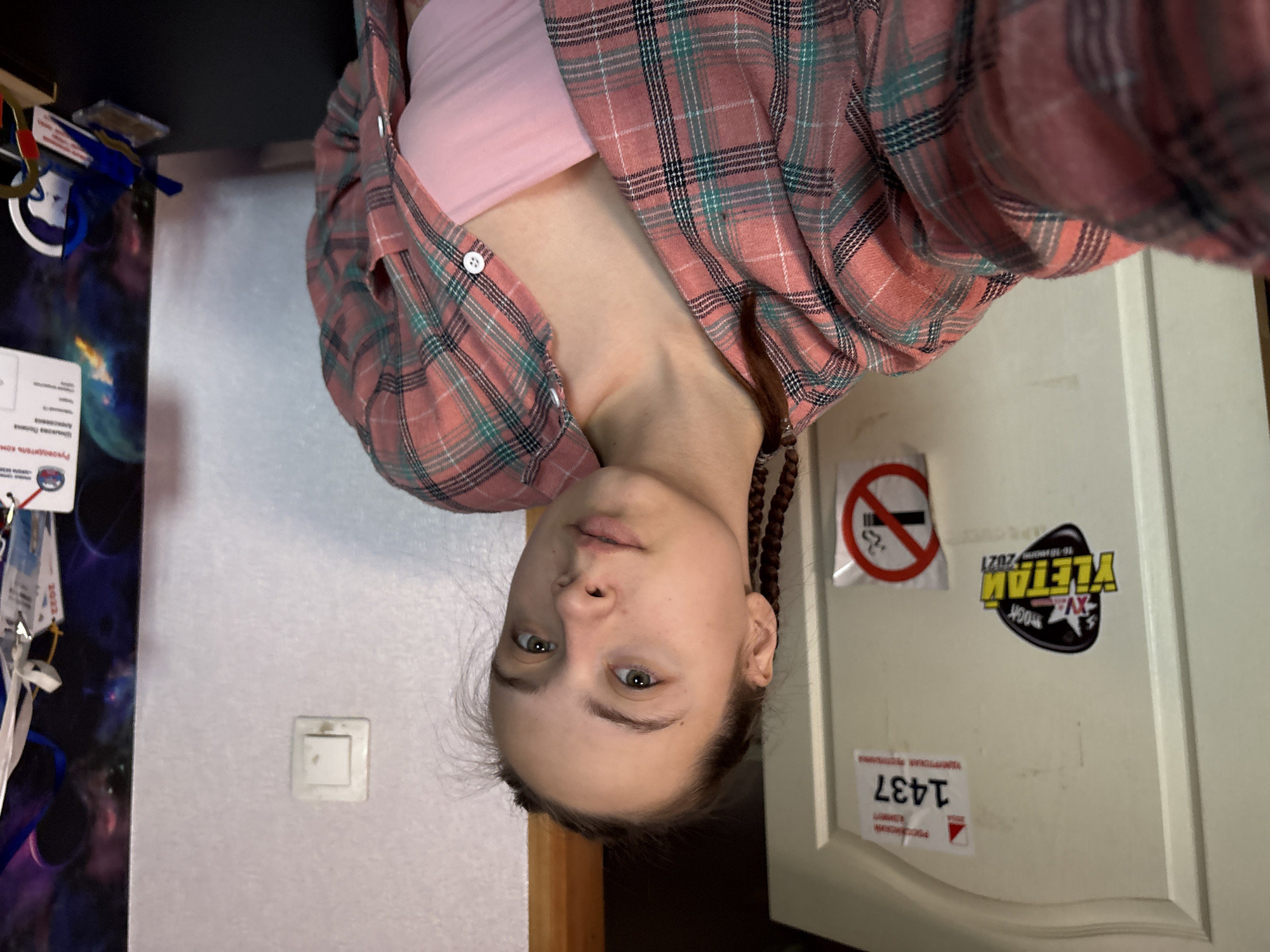 An indoor, close-up selfie shows a young person with light-colored eyes and braided dark hair, looking directly at the camera with a neutral expression. They are wearing a light pink crop top underneath an unbuttoned plaid shirt in shades of pink, green, and black. A wooden bead bracelet is visible on their right wrist.
The background on the left features a dark, patterned wall or poster depicting celestial bodies like planets and stars. Hanging items, possibly keys or lanyards, are visible near the top left corner. A white light switch is mounted on a plain white wall further to the right. On the far right, a white door is adorned with several stickers. These include a "No Smoking" symbol, a black and yellow sticker that reads "YLETAM XV ROCK 16-18 JULY 2021", and a white sticker with red text displaying "1437" above "Пермский край" (Perm Krai) and "Прикамье" (Prikamye), which corresponds to the given location context of Прикамский, Russia. The perspective suggests the camera is held above the person, capturing an upward-looking shot.