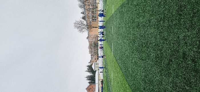 The image captures an outdoor football pitch in Pavlovsk, Russia, under an overcast, light grey sky. The foreground is dominated by a dark green artificial turf field, marked with white lines delineating playing areas. Patches of snow are visible along the edges of the pitch and on the ground beyond the playing surface. Approximately 15 to 20 individuals, primarily young males in athletic attire, are present on the field in the mid-ground. They are divided into two main groups based on their kits: one wearing blue tops and bottoms, and another wearing white tops with blue accents. One individual on the far right is depicted in the act of kicking a football. Others are standing or moving in various positions across the field, suggesting a training session or match. In the background, several multi-story buildings are visible, including a reddish-brown brick structure to the far left and a long, lighter-colored building further right. Bare deciduous trees and some evergreen trees are also present behind the buildings.
