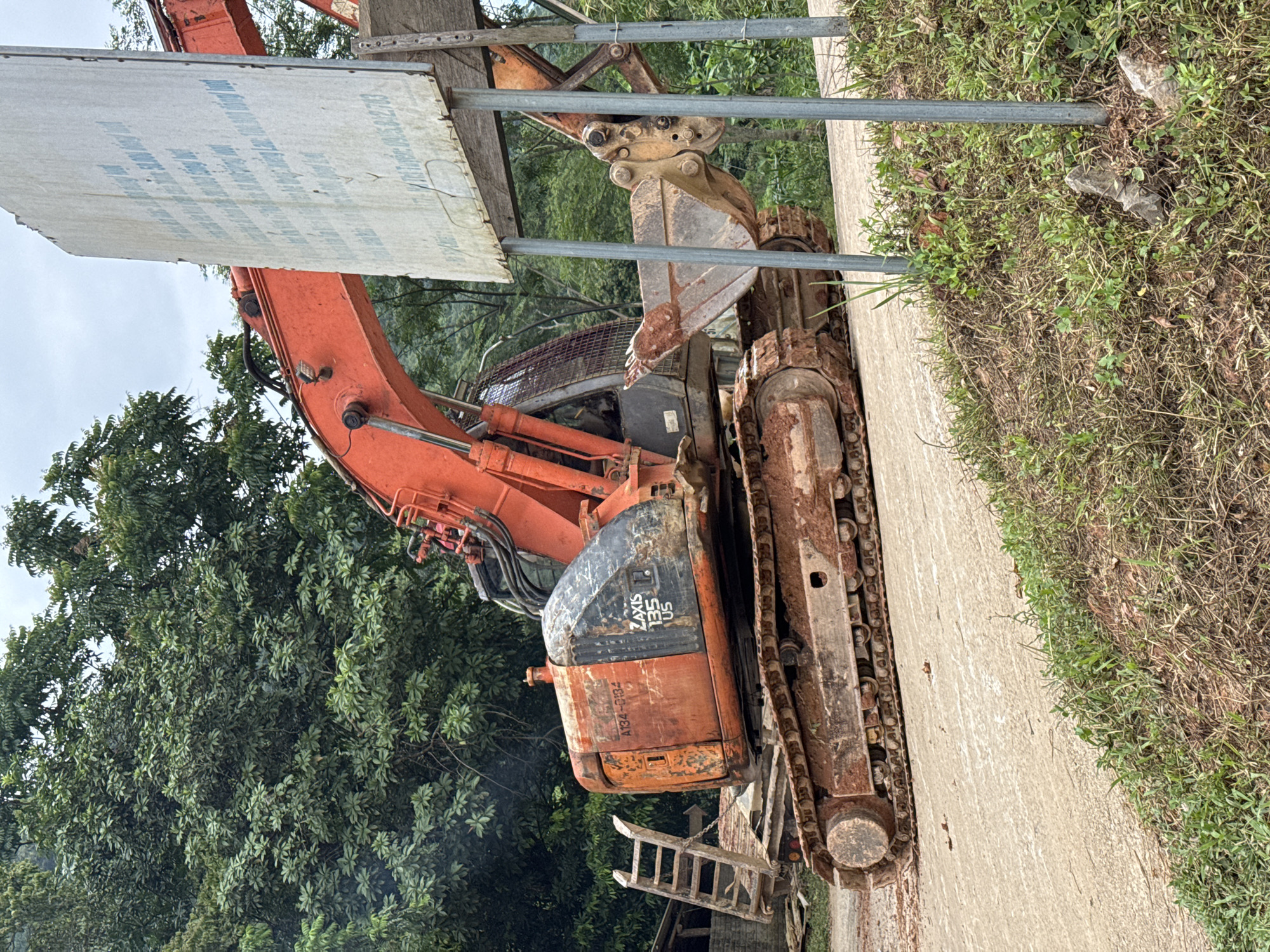 The image shows an excavator in a rural area. The excavator is orange and has the model name "ZAXIS 135 US" visible on its side. It is parked on a dirt road, and there is vegetation on either side of the road. In the background, there are trees and a cloudy sky. A large white sign with text in Vietnamese is also visible in the image. The location is identified as Cổ Lũng Commune, Vietnam. The scene appears to be a construction or excavation site. There are no people visible in the image.