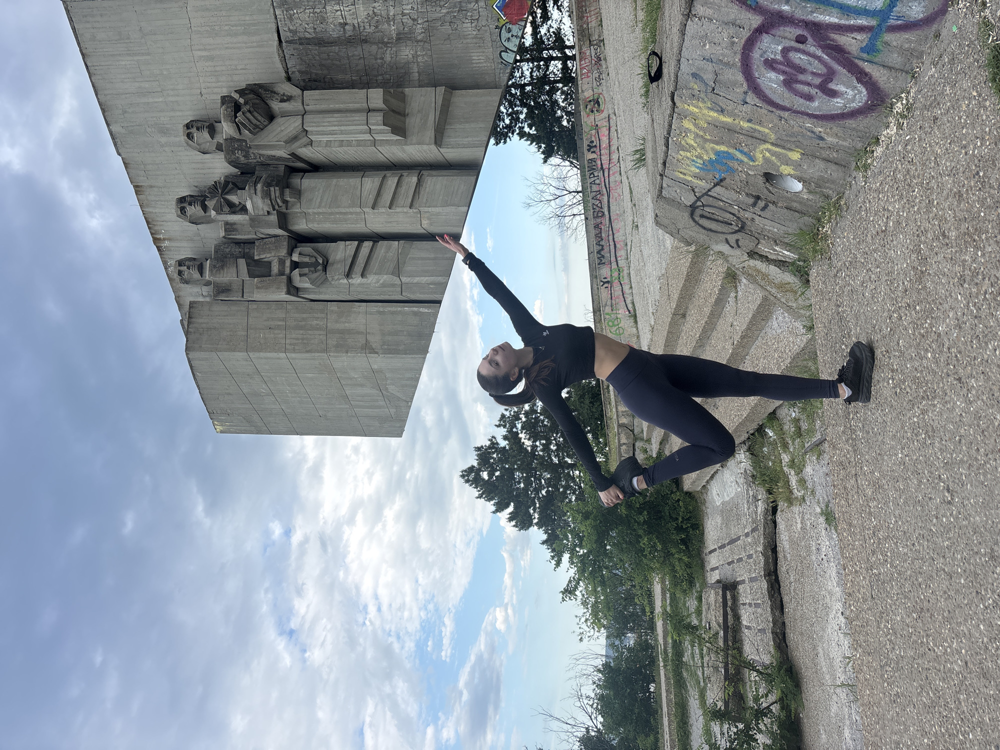 A woman in athletic wear performs a yoga pose in front of the Monument of the Bulgarian-Soviet Friendship in Varna, Bulgaria. The monument is concrete with socialist-era figures on it. The sky is visible between the monument and the ground. The woman is wearing a black crop top and leggings, with black shoes, and her hair pulled back. She is in a backbend with one arm raised towards the monument and the other reaching towards her foot. The ground is concrete with some graffiti. Vegetation is visible around the concrete structures.