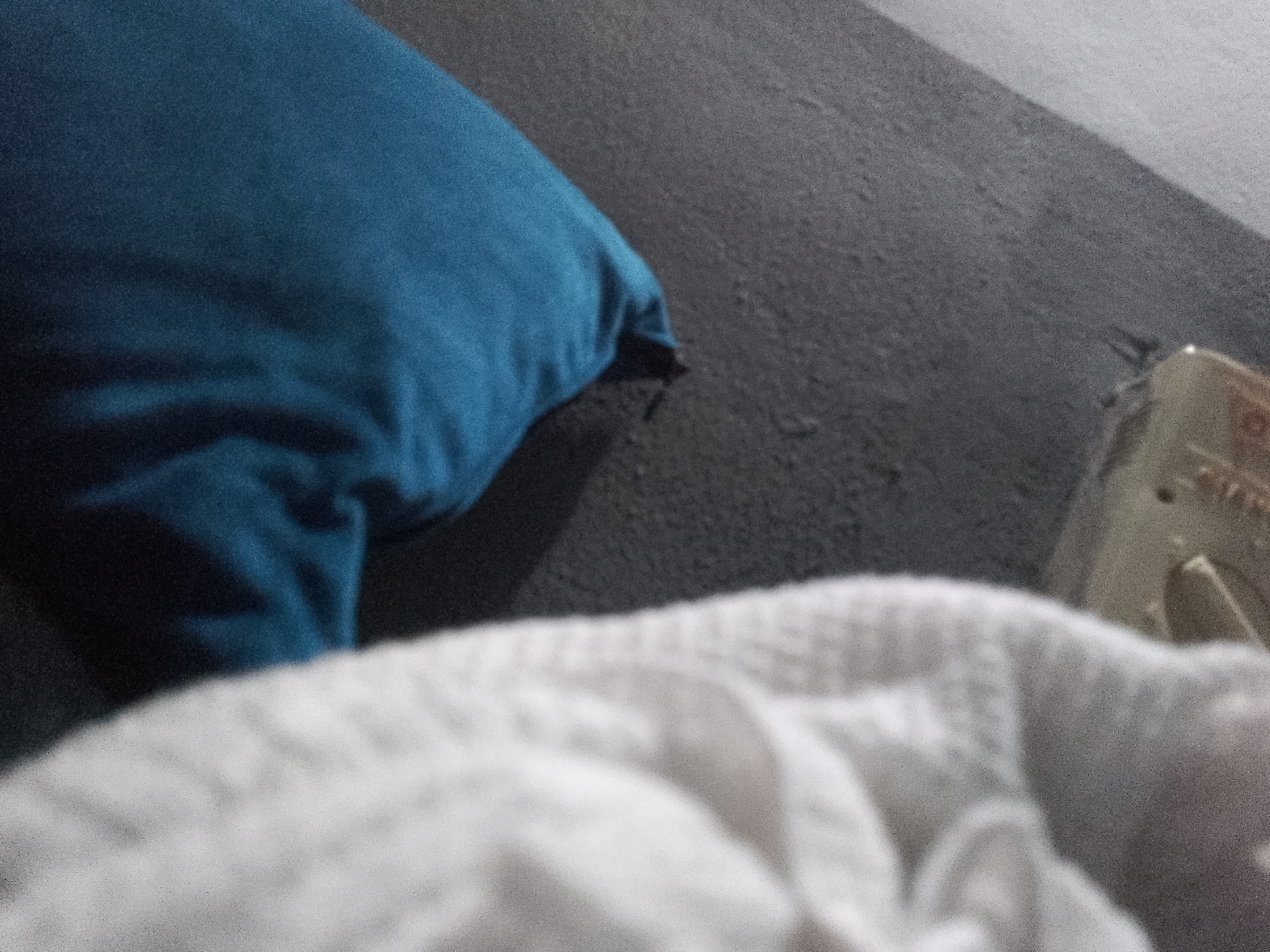 The image displays a close-up of a textured surface, likely a carpet or upholstery, with a blue cushion in the upper left quadrant. A white fabric, possibly a blanket or towel, fills the foreground. The lighting suggests an indoor setting. There are no discernible features to indicate the location in Jauro Yinu, Nigeria, or any specific actions, interactions, or people. The overall scene is static and lacks context.