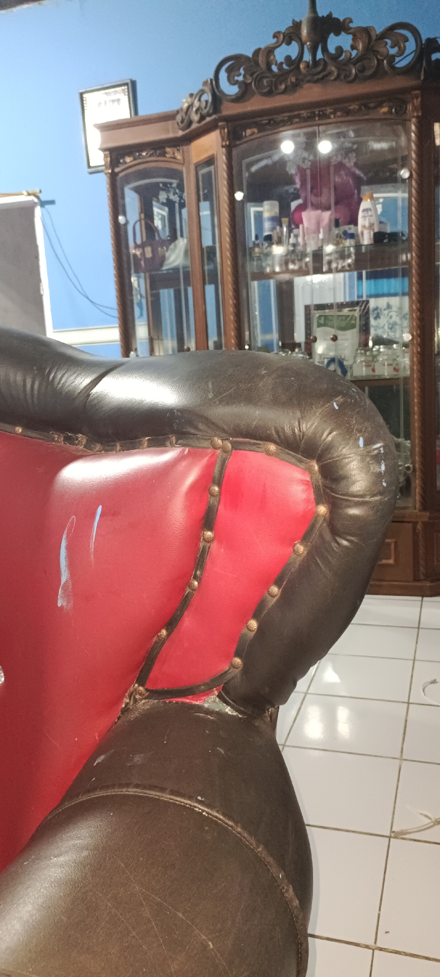 A close-up, low-angle shot shows the dark brown leather armrest of a chair in the foreground. The chair's inner cushion is a bright red, with decorative gold studs outlining the seam where the red meets the brown leather. A few faint blue smudges are visible on the red portion of the chair. In the background, a tall, dark wood display cabinet with glass doors stands against a bright blue wall. The cabinet contains various items, including bottles and decorative objects. The floor is tiled in white squares. The scene appears to be an indoor residential setting in Damuli Pekan, Indonesia.