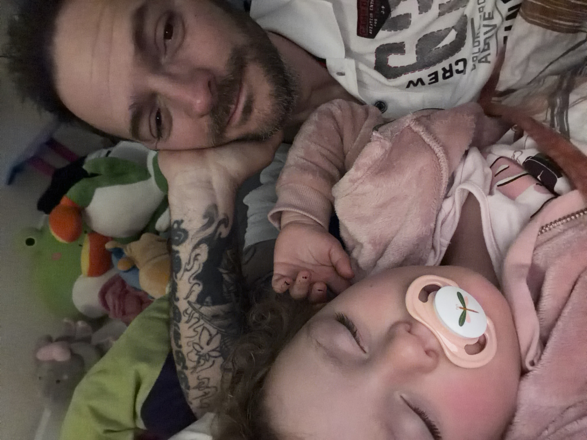 A man with a dark beard and tattoos on his left arm is lying next to a young child sleeping with a pacifier in their mouth. They appear to be lying on a bed surrounded by stuffed animals. The location is unknown and cannot be determined from the image.