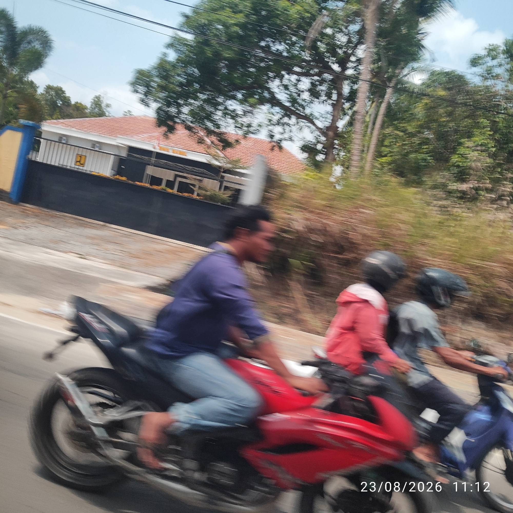 The image captures a motion-blurred scene of three individuals riding motorcycles on a road, likely in the city of Perawas, Indonesia. In the foreground, a person wearing a purple shirt and blue jeans rides a red motorcycle, with their left leg extended. Following closely behind, another person in a red jacket and dark pants is on a second motorcycle. Further back, a third individual in a grey shirt and dark pants is visible on a blue motorcycle. The environment includes a paved road with some gravel and debris. In the background, a building with a red-tiled roof and white walls is partially visible, bordered by a dark fence. Lush green trees and foliage line the right side of the road. The sky is a clear blue with some clouds. The timestamp "23/08/2026 11:12" is overlaid on the lower right portion of the image.