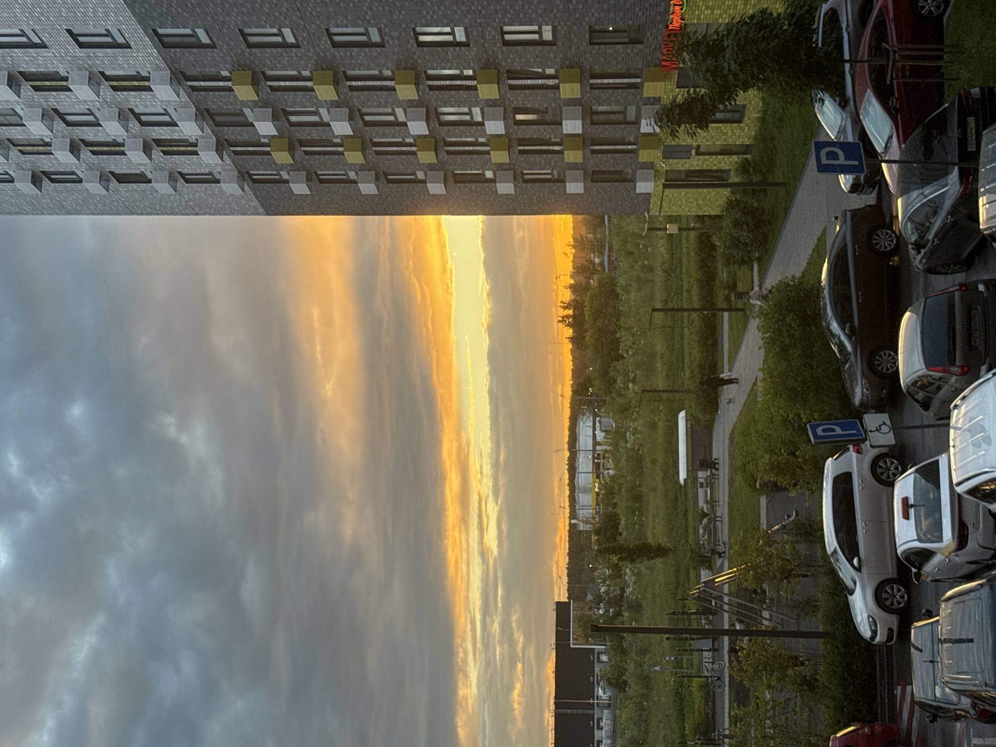 This is a vertical photograph of a residential building and a parking lot at sunset, likely taken in Drozhzhino, Russia. The building is modern, with a facade of gray brick and a repeating pattern of windows and balconies, some of which are accented with yellow. To the right of the building is a parking lot filled with various cars. One car is white, and another is dark colored, with a parking sign displaying a blue 'P' symbol visible above them. A small, pale-colored figure, possibly a person, can be seen walking on a paved path in the middle ground, near some green foliage. The sky is dramatic, with a striking contrast between the dark, brooding clouds on the left and the vibrant, fiery orange and yellow hues of the sunset on the right, indicating the time of day. The overall mood is tranquil, with the setting sun casting a warm glow over the scene. No specific event appears to be taking place, suggesting a typical evening in this suburban or semi-urban setting.