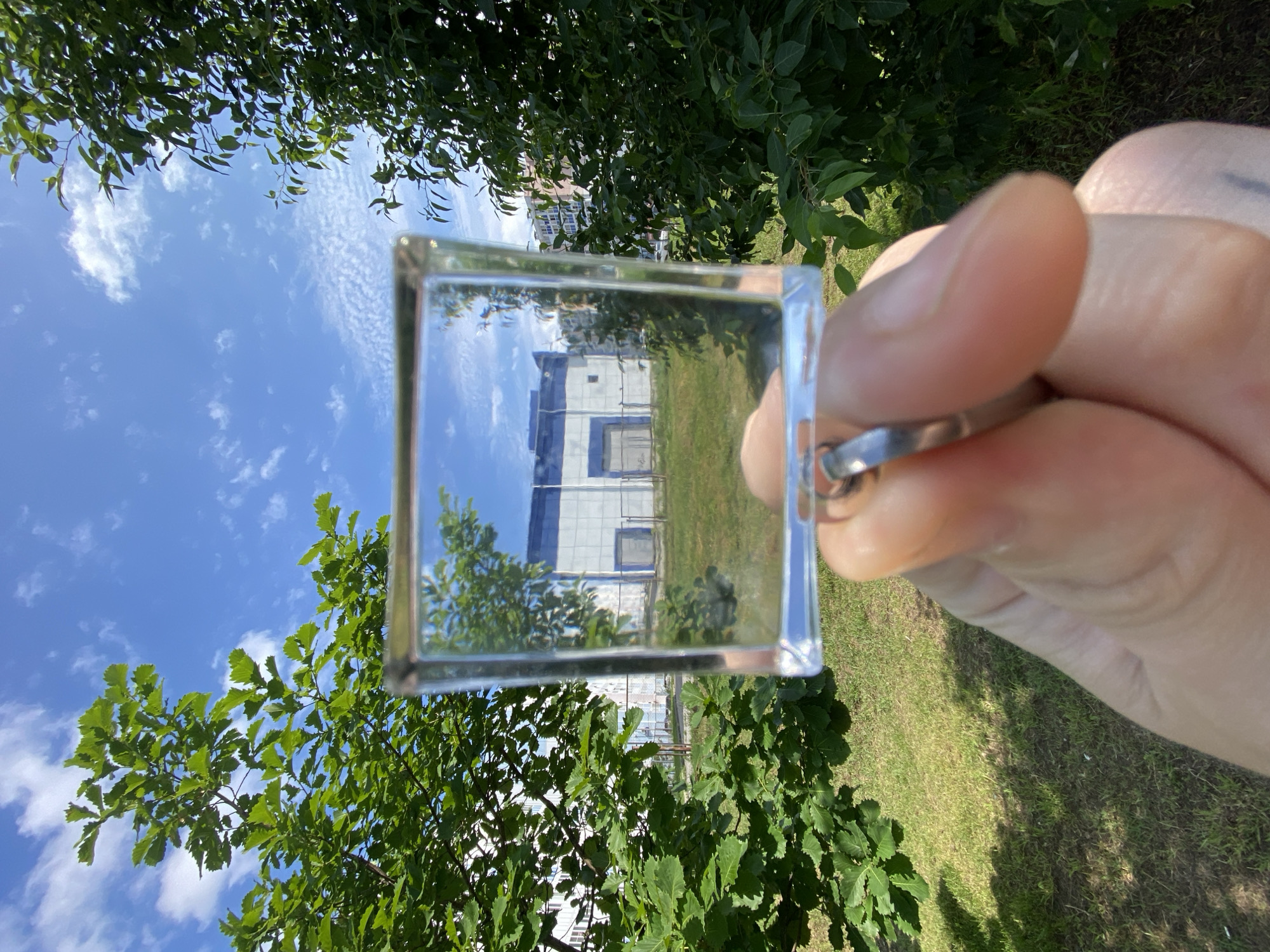 In Khabarovsk, Russia, a hand holds a clear, square glass object in the foreground, reflecting a building with white and blue panels and a large grey door. The building appears to be industrial or a warehouse, set against a backdrop of a bright blue sky with scattered white clouds. Green trees with lush foliage frame the scene on either side, with the leaves of one tree on the left being particularly vibrant and large. A patch of green grass covers the ground in front of the building. The image is a close-up, with the hand and the glass object being the primary subjects.