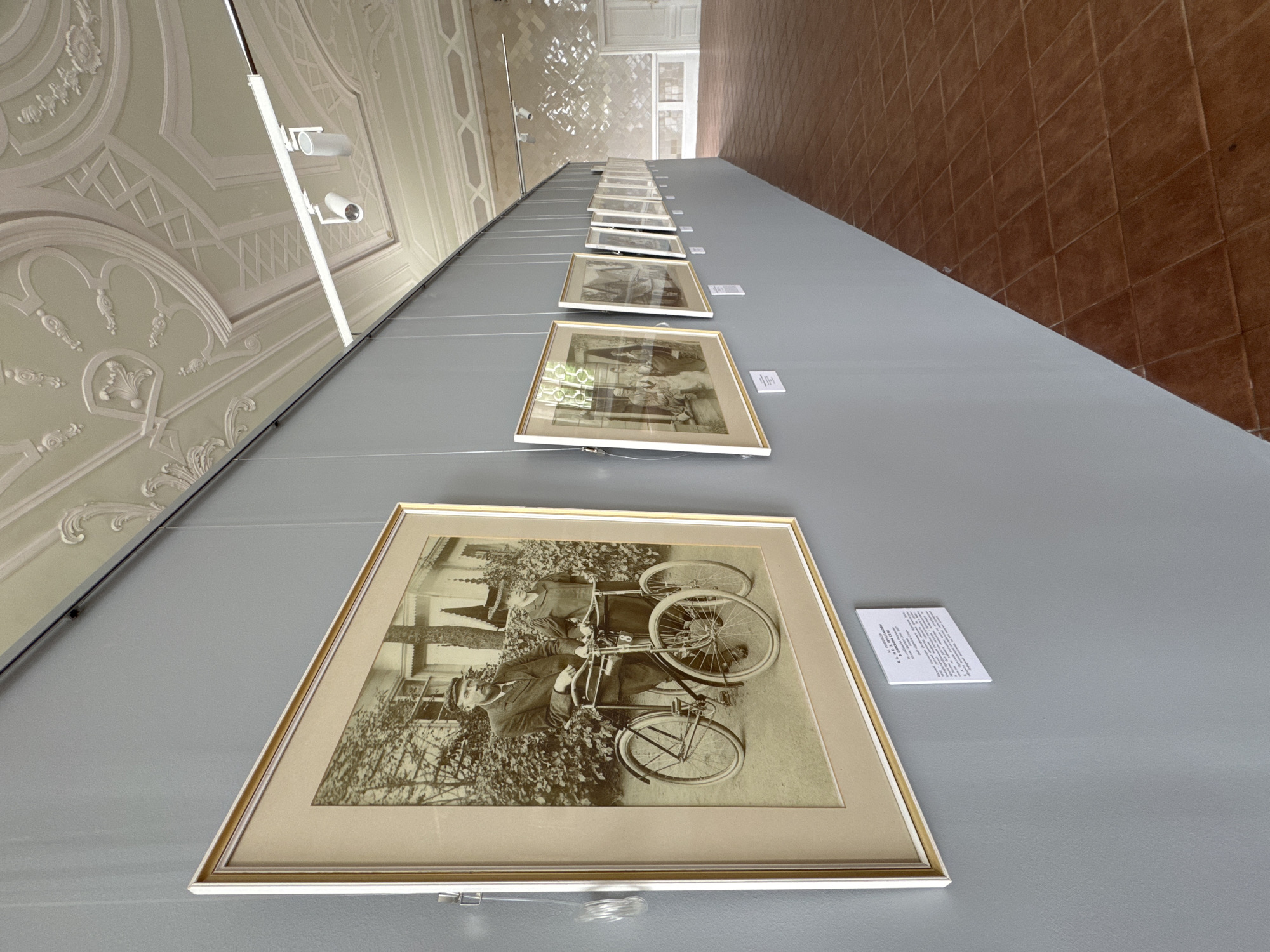 In the city of Pushkin, Russia, within a room featuring a decorative plaster ceiling, a row of framed black and white photographs is displayed on a gray wall. The closest photograph, prominently featured, shows a man and a woman standing beside two bicycles. The man, wearing a cap and suit, leans on the handlebars of one bicycle, while the woman, in a dress and hat, stands next to the other. Both bicycles appear to be vintage models. The photographs are hung from the ceiling by thin wires and are illuminated by track lighting. The room has a warm-toned tiled floor and the wall on the right side of the image is adorned with a textured pattern.