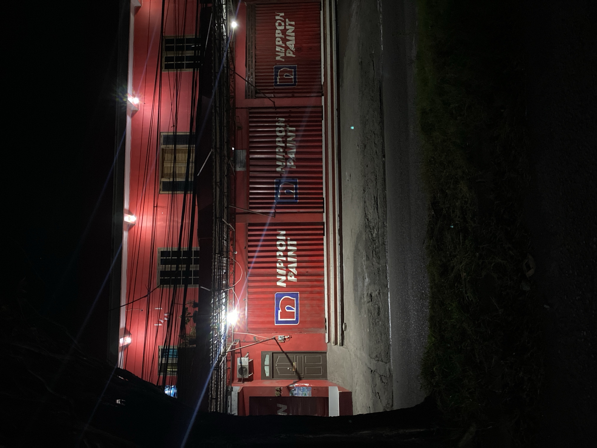 This is a nighttime, eye-level, medium shot of a red building with the "NIPPON PAINT" logo displayed on its metal roller shutters in Kuta Selatan, Indonesia. The building appears to be a storefront. The front of the building features several large, red metal roller shutters, two of which prominently display the "NIPPON PAINT" logo in white lettering. The logo includes a stylized white "n" within a blue square. To the left of the shutters, there is a brown wooden door and an air conditioning unit mounted on the wall. The building's upper facade is also red, with white trim and visible electrical wires strung across the front. On the upper level, there are windows with wooden frames and dark shutters. The area in front of the building is a paved street or sidewalk, with some grass and foliage to the left. The lighting is artificial, with spotlights illuminating the storefront. The scene is dark and appears to be deserted.