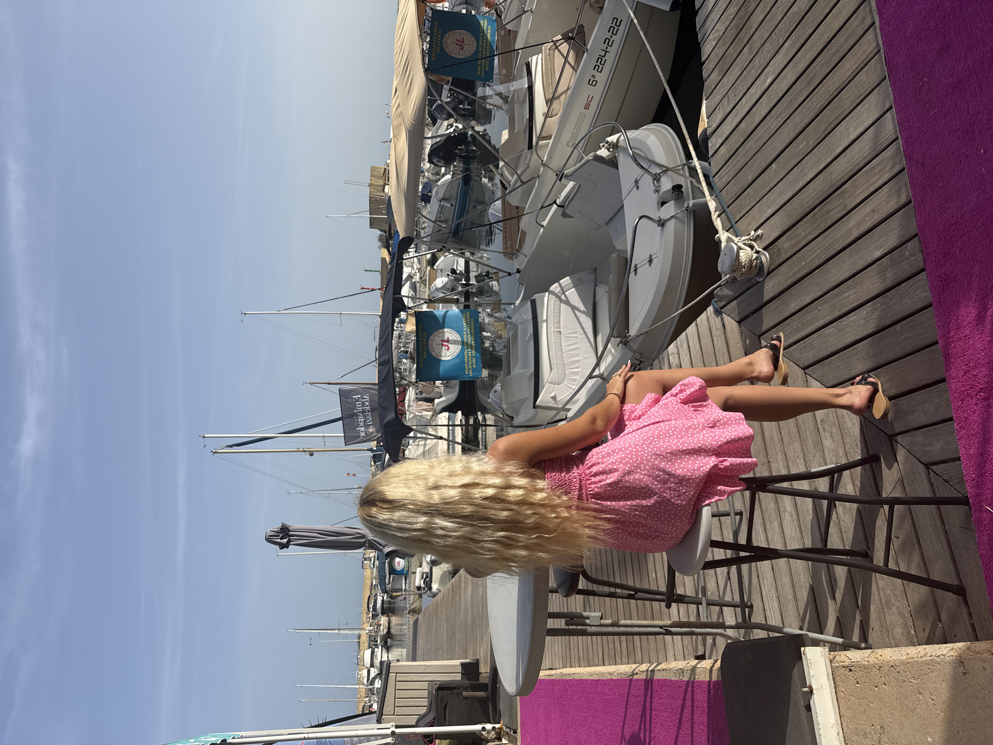 A woman with long blonde hair sits on a stool facing a marina. She is wearing a pink, polka-dot dress and sandals. She is looking out at a body of water with several boats docked. The boats are of various sizes and types, some are small motorboats and others appear to be sailboats. There are also several large umbrellas and signs visible in the marina. The scene is bathed in sunlight, and the sky is clear and blue. The ground is a wooden dock, and there is a table and a bright pink carpet in the foreground.