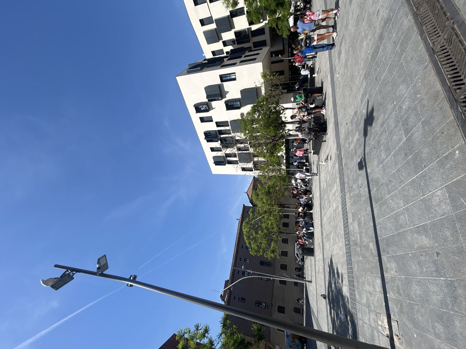 The image depicts a bright, sunny daytime scene in an urban plaza, identified as being in Barcelona, Spain. The sky is a vivid, clear blue, traversed by a few faint aircraft contrails.
On the right side of the frame, a tall, modern white building with numerous dark, recessed window frames or balconies dominates the skyline. In contrast, an older, darker building with many small windows and a reddish-brown tiled roof section is visible on the left. Green trees are interspersed along the pedestrian area, casting shadows.
The ground is paved with large, alternating light and dark gray tiles, featuring darker strips possibly for drainage. Long, distinct shadows of people, trees, and a tall lamppost stretch across the pavement, indicating strong, direct sunlight, likely during the mid-morning to late afternoon. A prominent dark metal lamppost with two modern light fixtures extends from the lower-left foreground into the sky.
Numerous people are present in the plaza, mostly gathered in relaxed groups. Many are seated along what appear to be benches or low ledges beneath the trees, while a few individuals are standing. They are dressed casually in warm-weather attire, suggesting they are enjoying the pleasant outdoor conditions. The overall activity appears to be casual relaxation and socializing in a public space.
No discernible text is visible anywhere in the image.