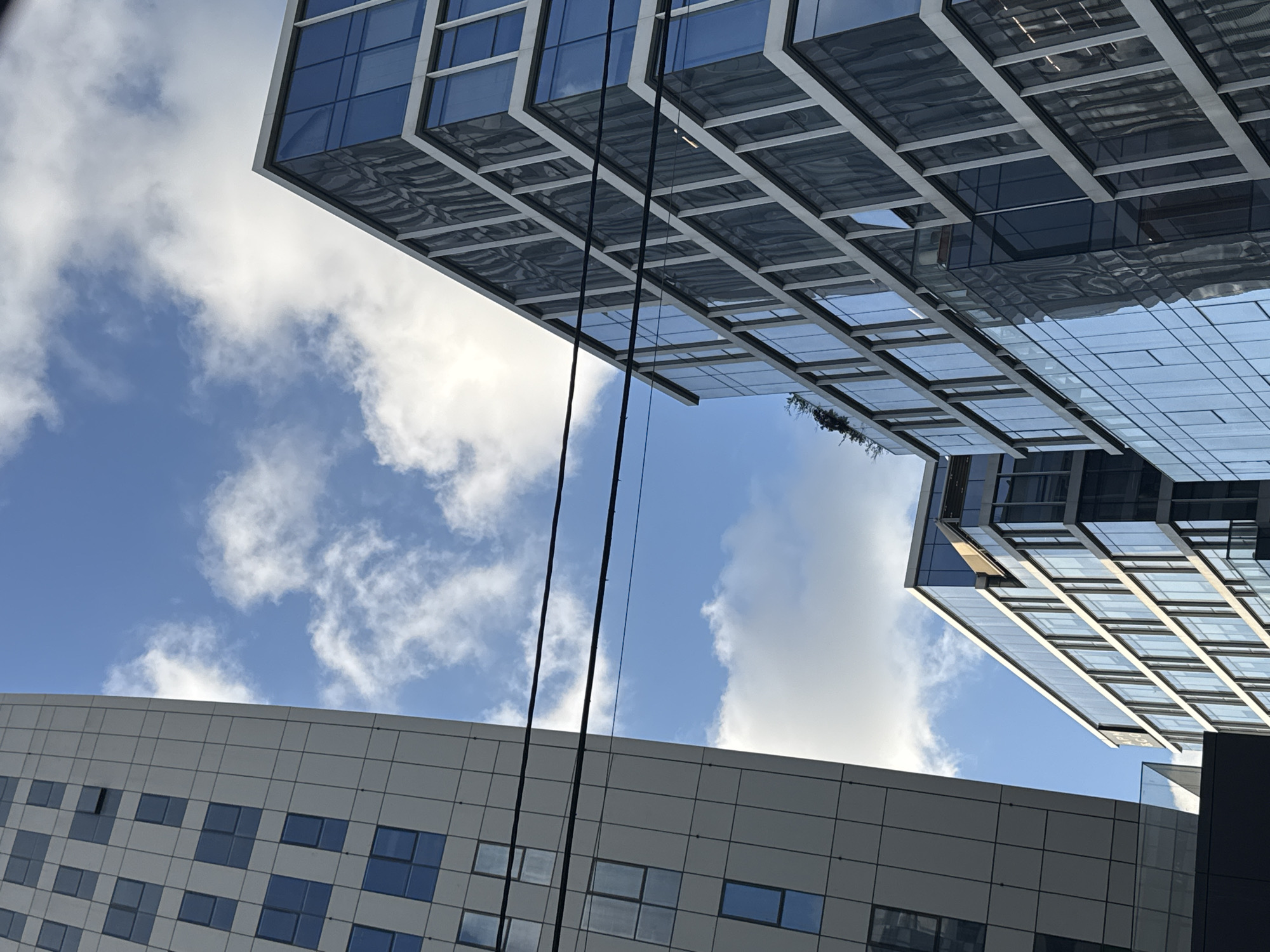 The image depicts a low-angle view of modern architecture against a blue sky with scattered clouds. The bottom of the frame shows a building with a curved facade and a grid pattern of windows. Above, two contemporary glass and steel structures angle towards each other, reflecting the sky. Three dark cables stretch between the buildings and the bottom of the frame. The image suggests an urban setting, potentially Tel Aviv, Israel, given its architectural style.