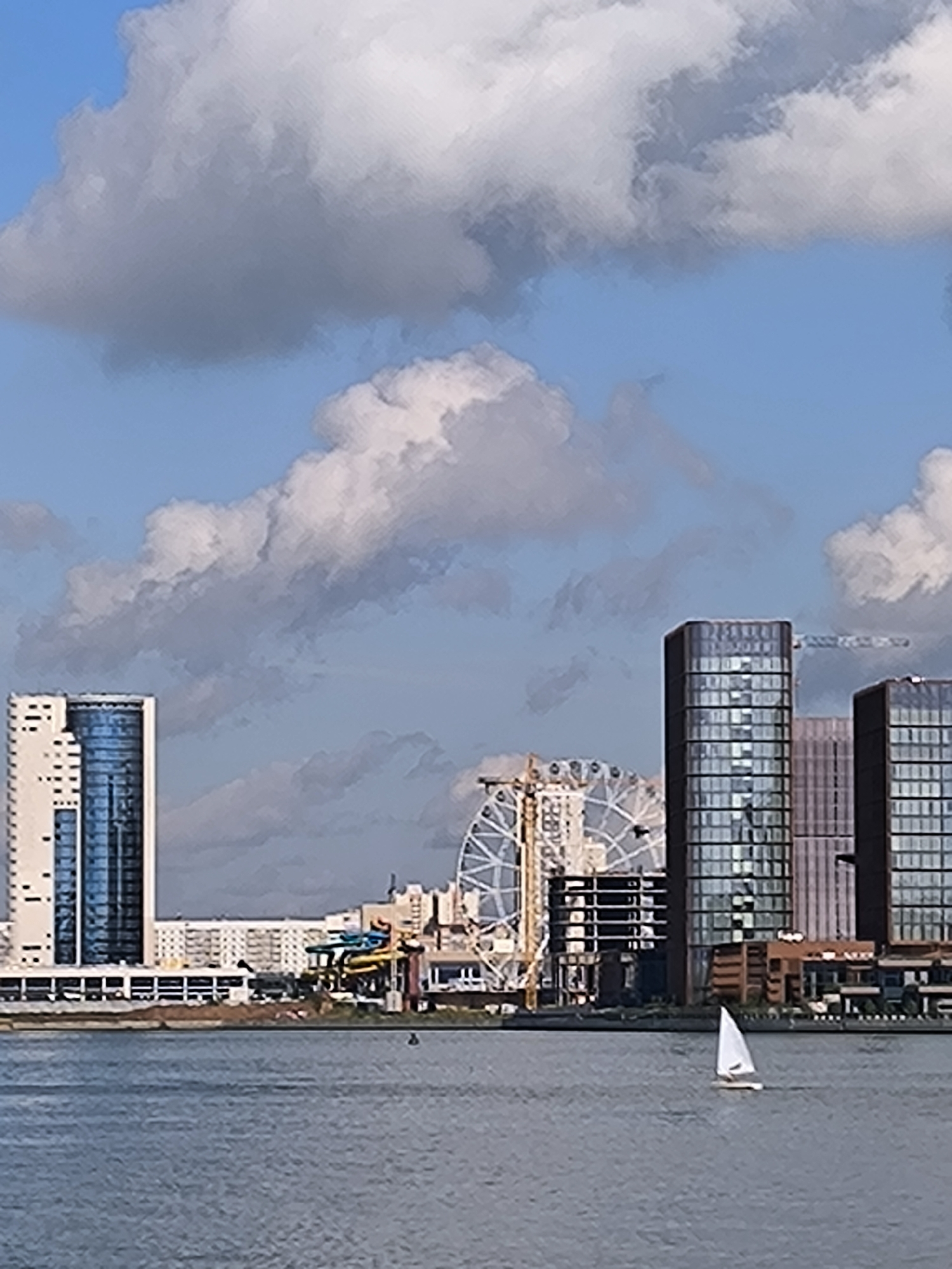 The image depicts a wide view of the Kazanka River in Kazan, Russia, on a sunny day with scattered clouds. On the river's edge, a modern cityscape unfolds, dominated by tall, glass-fronted buildings and a Ferris wheel. A small white sailboat with a single sail navigates the water in the foreground. The cityscape includes high-rise residential buildings, commercial structures, and construction cranes, indicating ongoing development. The water appears calm, reflecting the sky and buildings. The overall impression is one of a vibrant, developing urban waterfront.