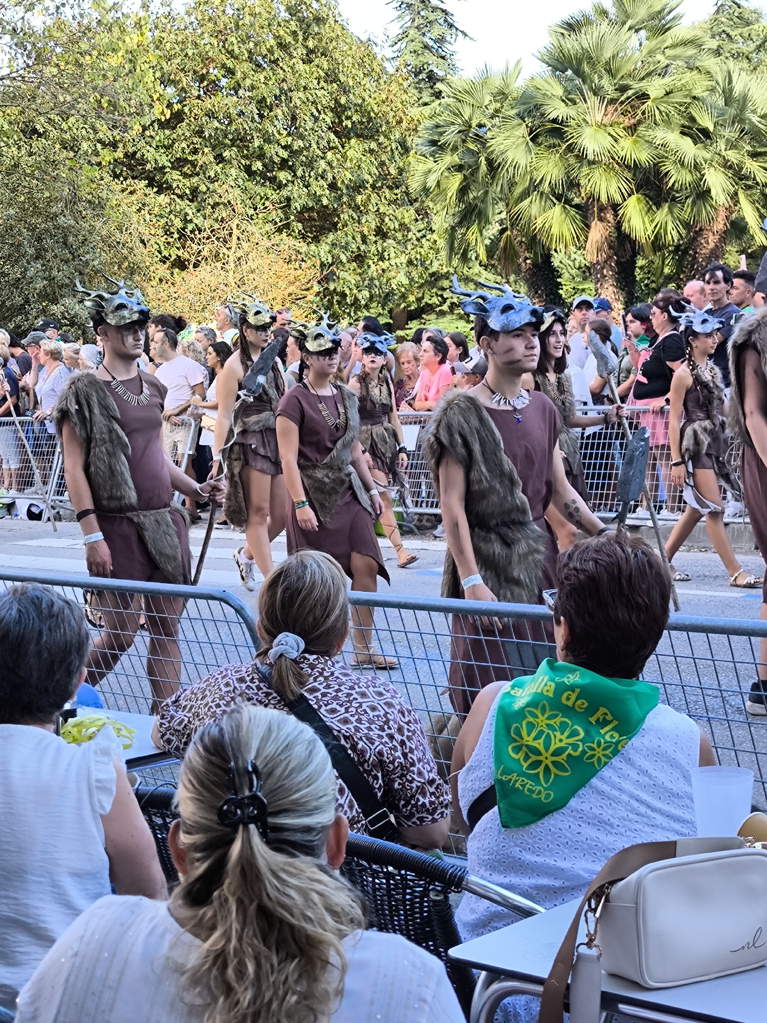In Laredo, Spain, a parade is underway, with participants dressed in historical costumes walking in a procession. Several individuals, appearing to be young adults, are in the foreground, wearing rustic, animalistic masks and garments made of fur and brown fabric, suggesting a historical or fantasy theme. They carry wooden staffs. Behind them, a crowd of spectators is visible, separated from the parade by a metal fence. Palm trees and leafy trees form the backdrop, indicating a warm climate. The scene appears to be a public event or festival.
