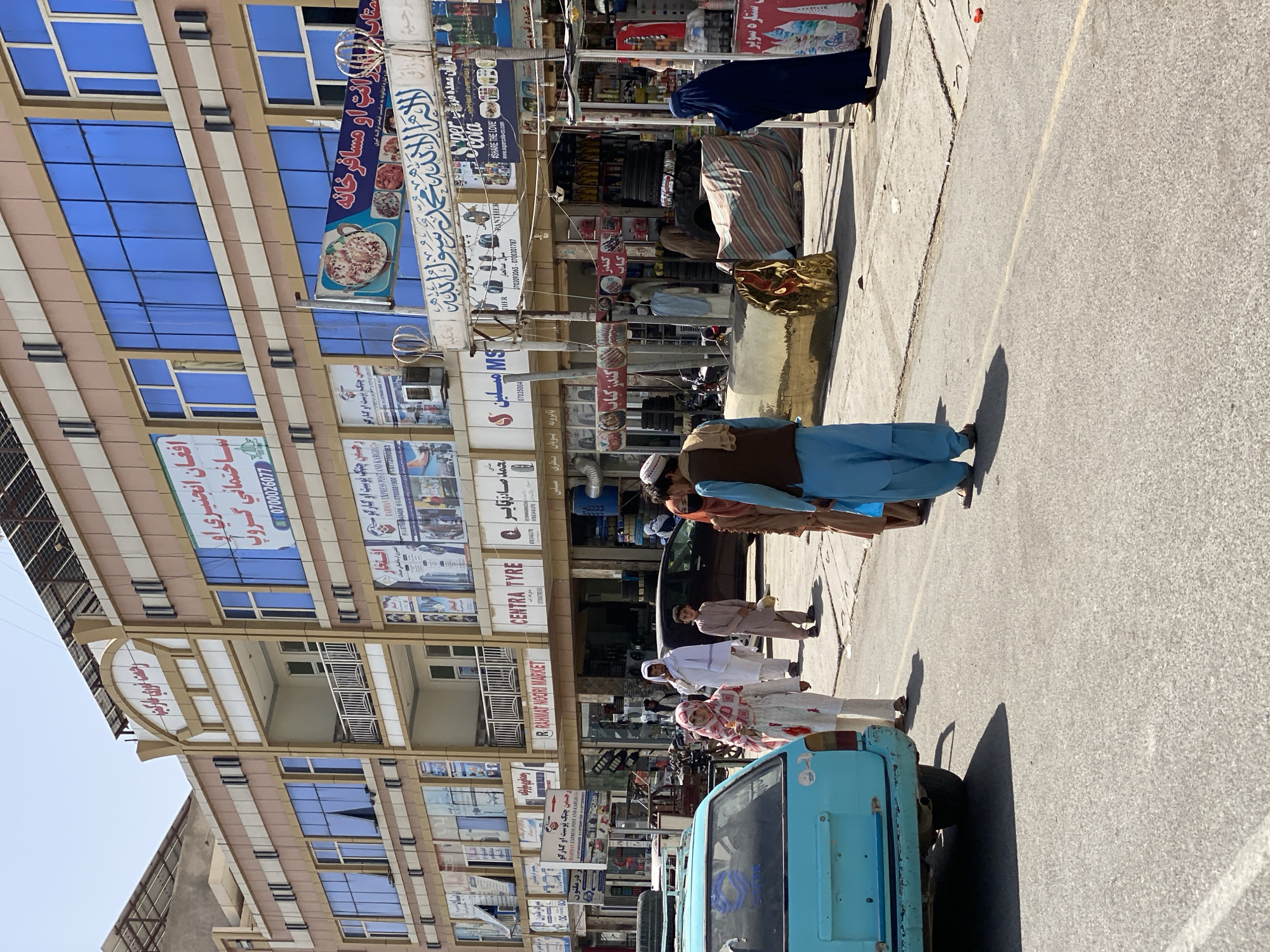 In this image taken in the city of Kandahar, Afghanistan, a bright blue car is parked on the right side of a paved street. Several people are walking on the sidewalk in front of a multi-story building with a beige and brown facade and large blue windows. A sign on the building reads "Rahmat Noori Market." Some of the people are dressed in traditional Afghan clothing. The building appears to be a market or commercial complex with various shops and advertisements on its facade. The sky is clear and the overall impression is a busy urban street scene in Afghanistan.