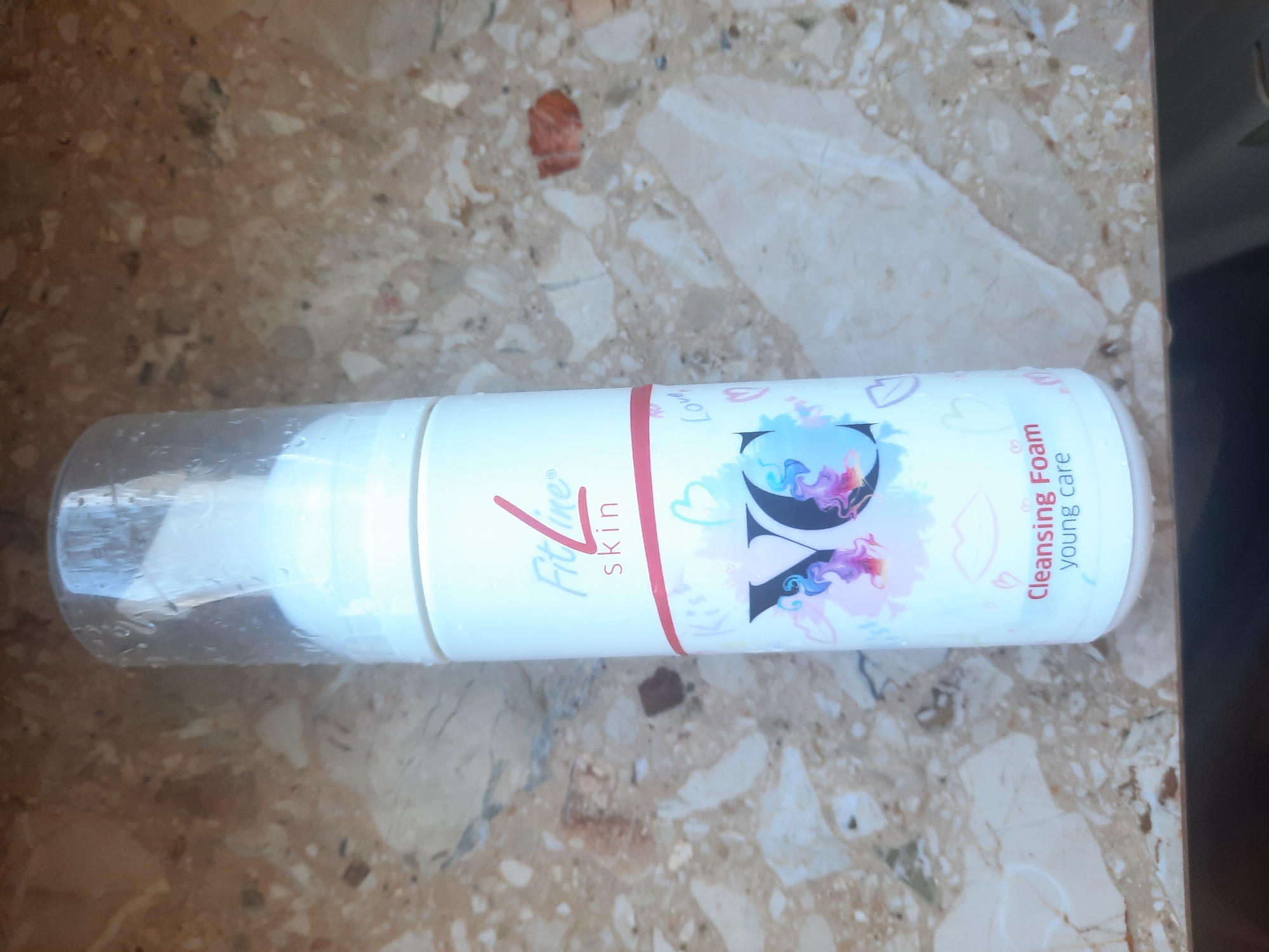 An overhead shot captures a white cylindrical bottle of FitLine skin cleansing foam, its cap slightly fogged. The bottle is positioned on a speckled brown and beige surface that resembles a marble countertop. The label on the bottle features the FitLine logo in red and blue, with the word "skin" beneath it in red text. A pink horizontal line separates this from the lower portion of the label, which displays the letters "YC" in large, stylized font, adorned with colorful abstract designs and small heart and lip motifs. Below "YC," the words "Cleansing Foam" appear in red, followed by "young care" in blue. The lighting suggests an indoor setting with diffused light.