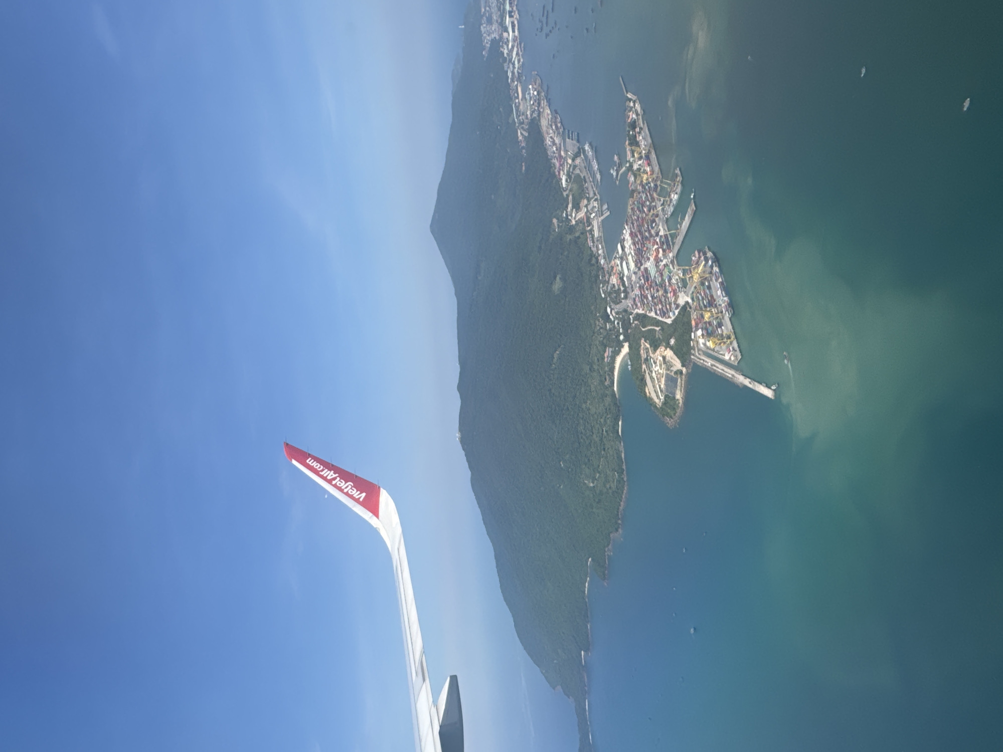 The image depicts an aerial view from an airplane, with a Vietjet Air wing visible in the lower left corner. In the distance, the city of Đà Nẵng, Vietnam is seen, specifically the Tiên Sa seaport on the Sơn Trà peninsula. The peninsula is covered in lush greenery. The seaport has numerous cranes, docks, and container storage areas. The sea surrounding the peninsula is a mix of green and blue, with several boats visible. The sky is clear and blue.