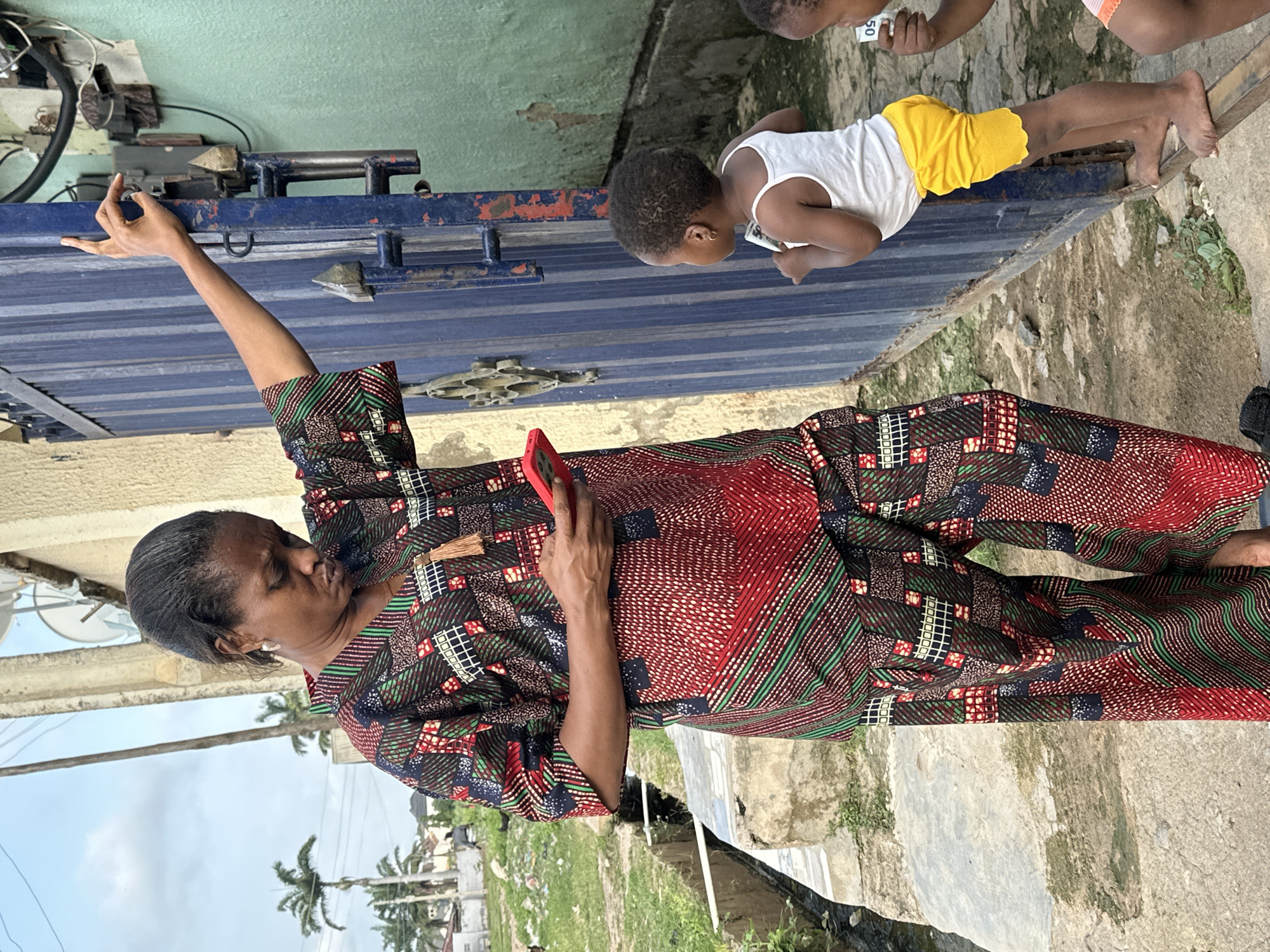 An adult woman, dressed in a patterned traditional African outfit, stands next to a blue metal gate, holding a red smartphone. She is looking down at the phone. Beside her, a young child wearing a white tank top and yellow shorts stands on a metal bar, peering through the gate. The environment appears to be an outdoor setting in Narayi, Nigeria, with an unpaved area and a drainage channel visible. The scene captures a moment of everyday life, possibly involving communication or observation.