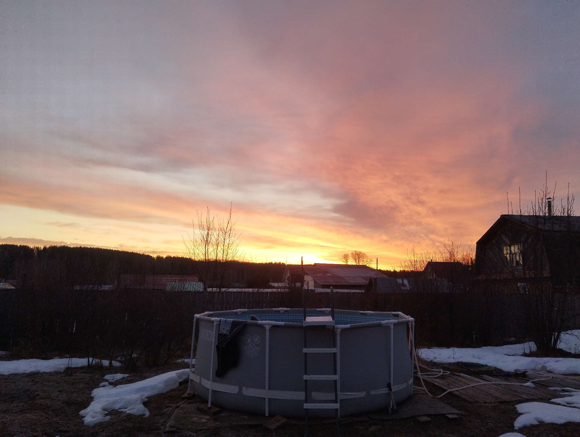 The image depicts an above-ground Intex swimming pool with a ladder attached, located in what appears to be a backyard or outdoor area in Grishanki, Russia. The pool is surrounded by patches of snow and dormant vegetation. A wooden structure partially covered by hoses is near the pool.
In the background, there are silhouettes of trees, fences, and buildings, including a house with a lit window. The sky is a vivid mix of pink, orange, and yellow hues, indicating either sunrise or sunset. There are no people visible in the image. The overall impression is of a transitional season, with lingering snow alongside signs of warmer weather.