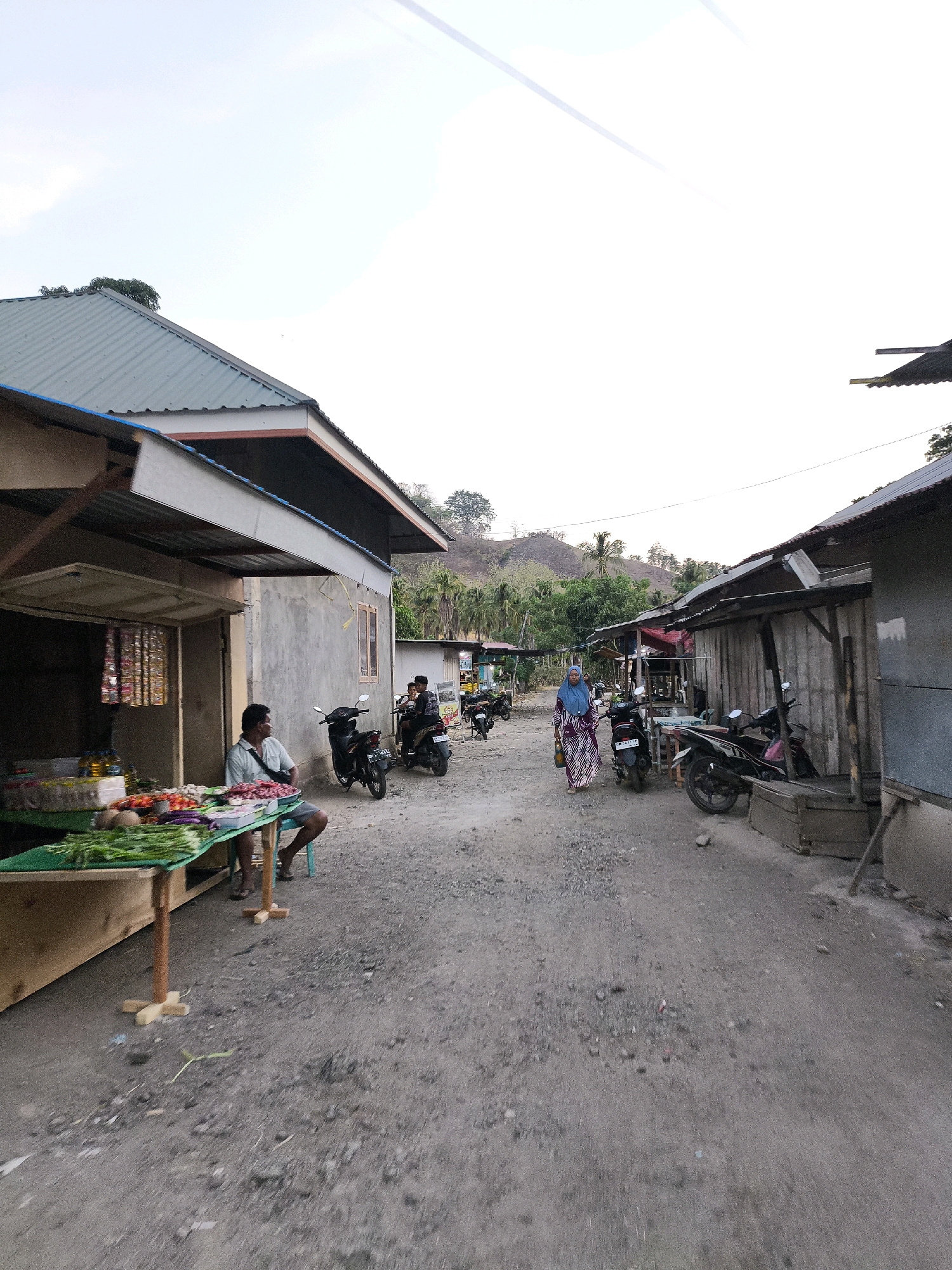 The image depicts a street scene in a rural village, possibly in Indonesia. A dirt road runs through the center of the frame, with buildings lining both sides. On the left, a small market stall displays fresh produce including vegetables and fruits. A man is seated on a stool at the stall. Several motorcycles are parked along the road and in front of the buildings. Two individuals are walking down the road, one wearing a blue hijab and a patterned dress, and another person on a motorcycle. In the background, hills covered with vegetation rise. The sky is overcast with some streaks of light. The overall impression is of a simple, everyday setting in a developing area.