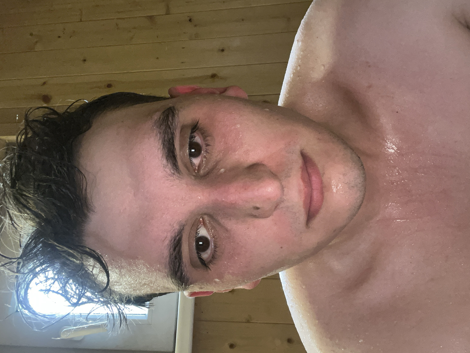 A close-up selfie shows a light-skinned person with wet, dark hair and brown eyes. The person's face and upper chest are covered in water droplets, suggesting sweat or recent washing. The background consists of wooden planks, indicating a sauna or similarly constructed room. A window is visible at the left side, with a bright light shining through it. Vasilyevo, Russia, is not identifiable from the image.