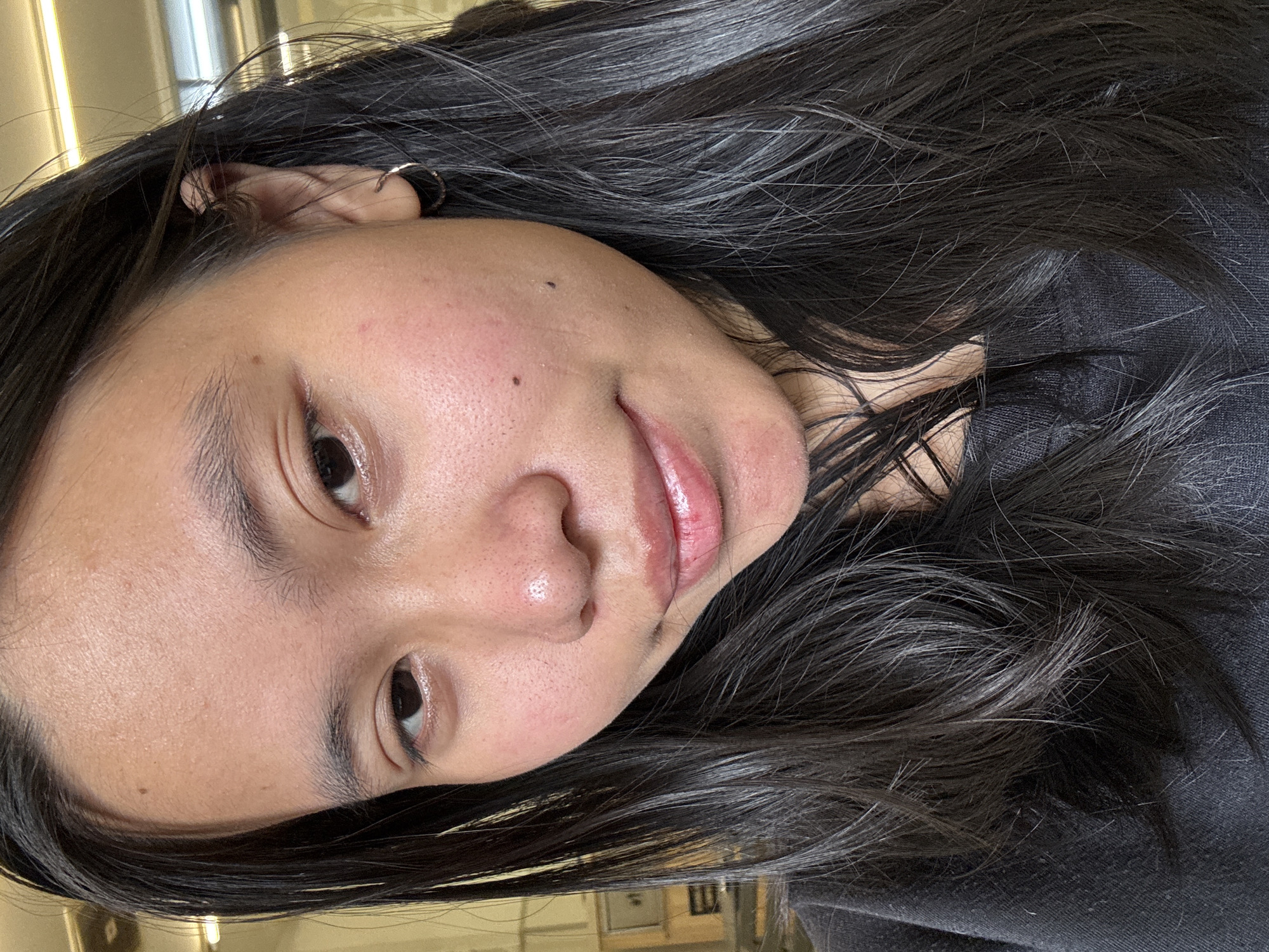A close-up, head-and-shoulders selfie of a young woman with long, dark hair and fair skin, taken indoors. She is wearing a dark t-shirt. Her face is turned slightly to her right, and she is looking towards the camera with a neutral expression. Her skin is slightly flushed on her cheeks and nose. A small hoop earring is visible in her right ear. The background is blurred and appears to be an indoor setting with some shelving units visible to the left. The image does not contain information about the location in East Jakarta, Indonesia.