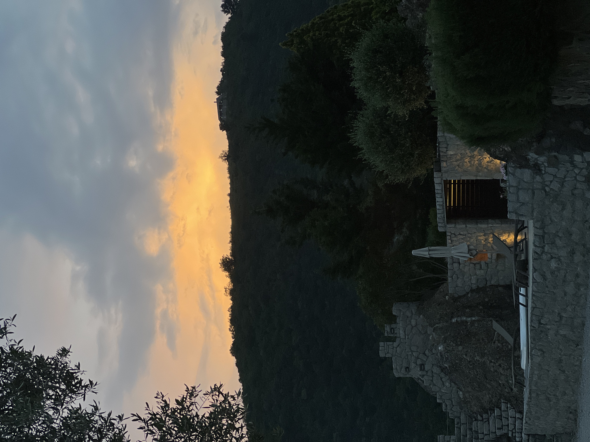 The image depicts an outdoor scene at dusk in Ivanovići, Montenegro. A hillside covered in dark green foliage rises in the center of the frame, silhouetted against a sky with streaks of yellow and gray clouds. To the right, a stone structure with a partially visible window is nestled amongst trees. A patio area with furniture and an umbrella are also visible. In the foreground, leafy branches extend into the frame on the lower left and right. The overall scene suggests a tranquil, mountainous location at twilight.