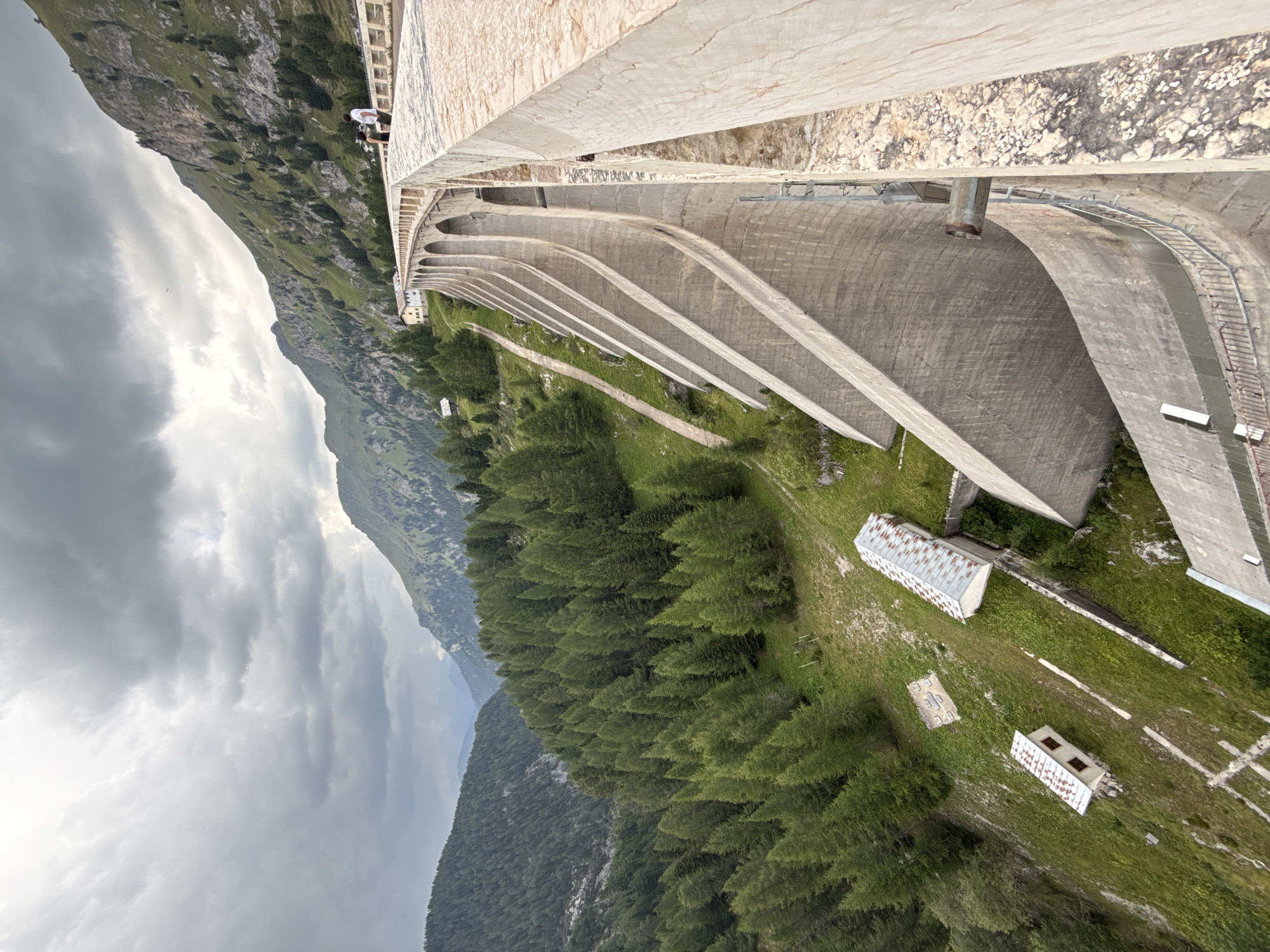 An extreme wide shot shows the large concrete dam of the Vajont Dam, located in the municipality of Canazei, Italy. The dam is a curved structure with arched openings. Two people stand on top of the dam, observing the mountainous landscape. The surrounding environment is characterized by steep, green mountains covered in trees, with a cloudy sky overhead. In the foreground, the dam's massive concrete structure dominates the scene. Smaller buildings with corrugated roofs are visible on the grassy slope below the dam. The image captures the scale and grandeur of the dam and its natural surroundings.