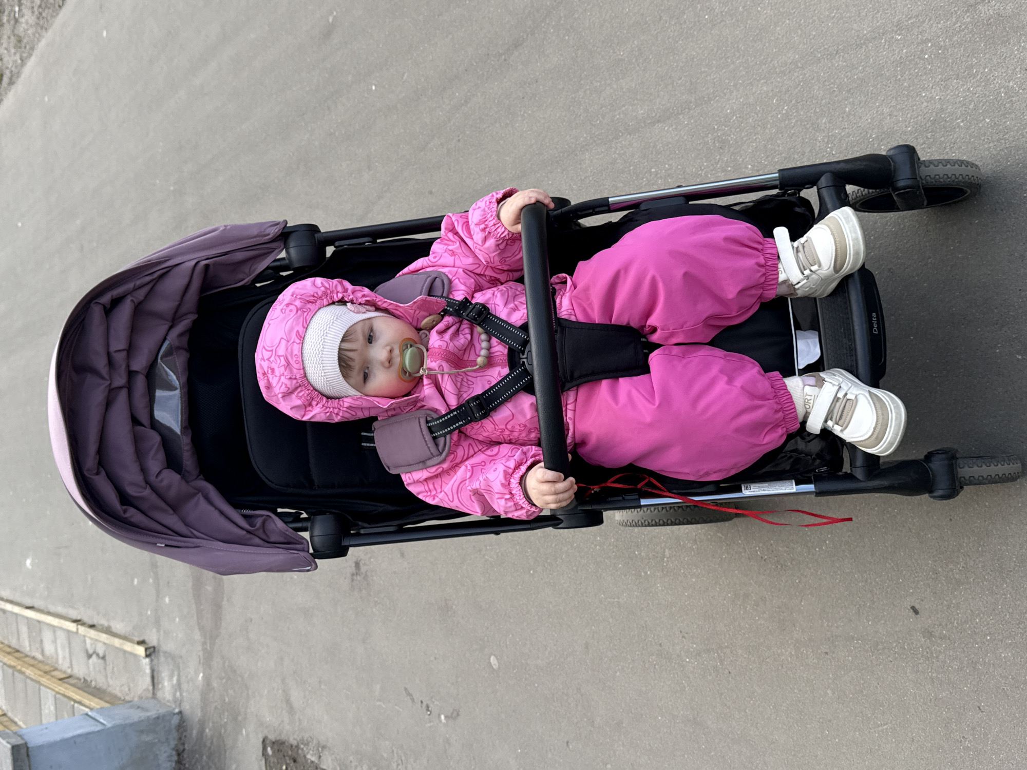 A medium-angle shot captures a baby in a pink snowsuit inside a black and purple stroller, situated on a light gray concrete surface. The baby wears a white hat and white shoes, and has a pacifier in its mouth. The stroller is positioned on a paved area that appears to be a sidewalk or walkway. No signage or landmarks indicating the specific location in Moscow, Russia, are visible.