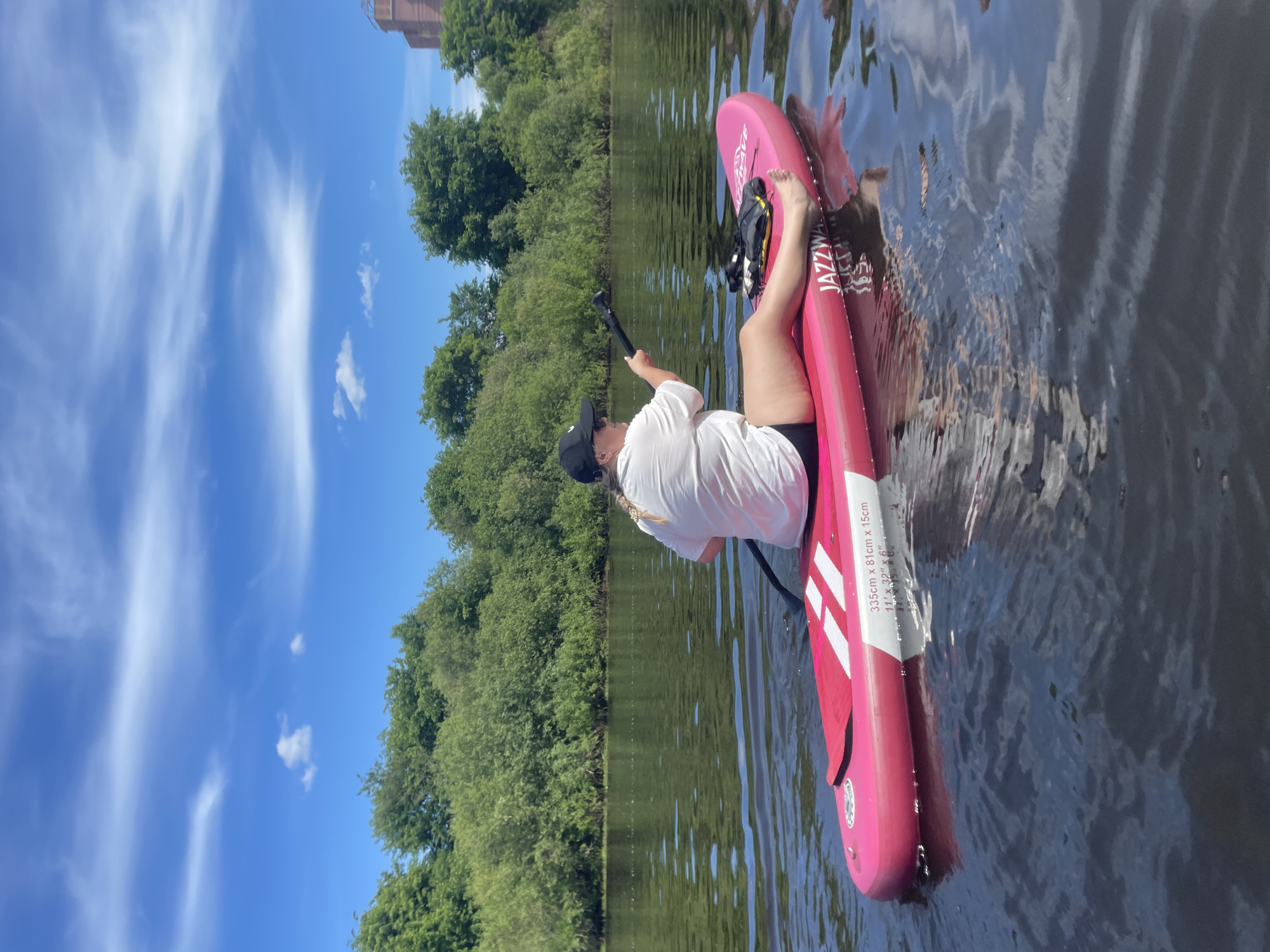 A woman is paddleboarding on a body of water in the city of Vologda, Russia, under a clear blue sky.
The woman, with light-colored hair styled in a braid and wearing a black baseball cap, a white t-shirt, and black shorts, sits on a bright pink inflatable paddleboard. She holds a black paddle, positioned mid-stroke, facing slightly to her left. Her paddleboard, a "JAZZY WAVE" model, displays dimensions "335cm x 81cm x 15cm" (11' x 32'' x 6''). A black and yellow pack or life vest rests on the board near her bare feet.
The dark water reflects the sky and shows numerous ripples. The far bank is densely covered with green deciduous trees and bushes. Above the treeline on the right side of the image, a distinctive reddish-brown building with a prominent upper structure is partially visible, characteristic of the Vologda cityscape.
The sky above is bright blue with scattered white cirrus and cumulus clouds. The scene is illuminated by strong sunlight, suggesting a clear summer day.